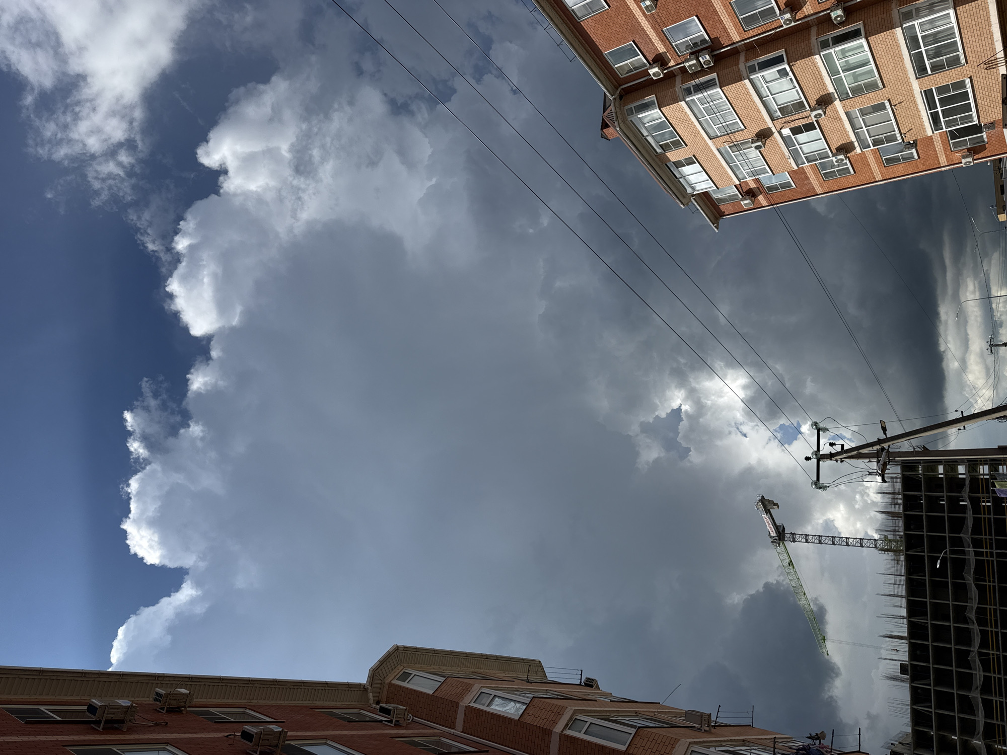 The image is taken from a low angle, looking upwards towards the sky, which is dominated by a large, billowing storm cloud. Patches of blue sky are visible on the left side of the frame. The cloudscape is framed by buildings on three sides. At the top is a section of a brick building with multiple windows and air conditioning units. On the right side of the frame, a construction site is visible, including a crane and the skeletal structure of a tall building. At the bottom, there is a portion of another brick building. Power lines stretch across the sky. The location appears to be an urban area.