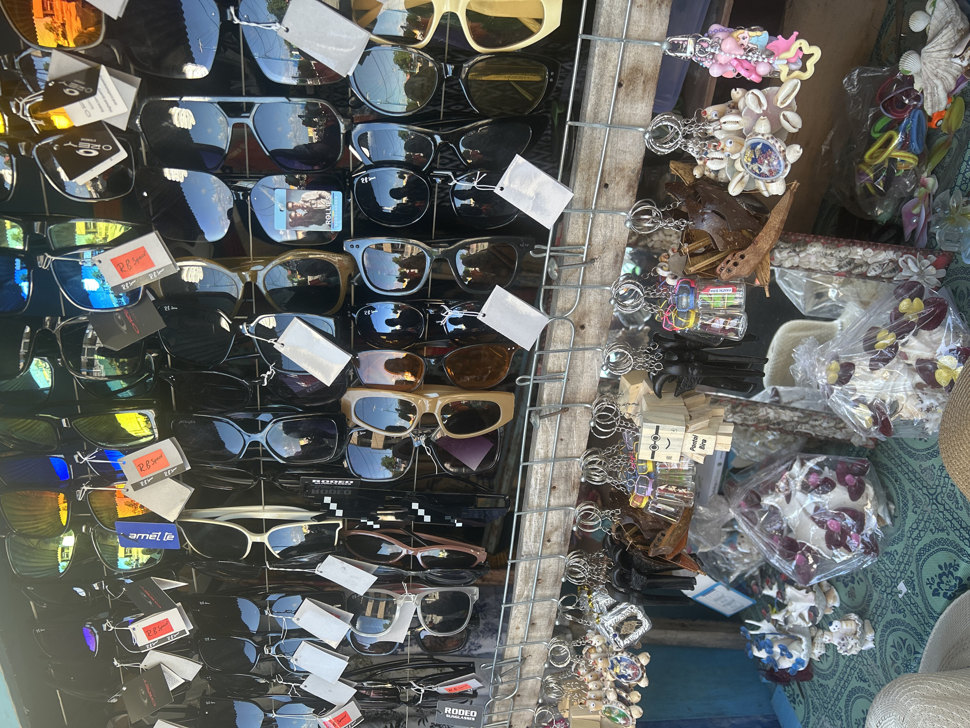 A close-up, eye-level shot captures a stall displaying a variety of sunglasses and keychains. The sunglasses, predominantly black and featuring tinted lenses, are arranged in rows on a metal rack. Some are adorned with price tags and brand labels such as "OAKLEY," "RB Sport," and "RODEO." Below the sunglasses, numerous keychains hang from hooks. These keychains are diverse, featuring shell decorations, wooden carvings, miniature figures, and colorful plastic charms. The stall is set against a backdrop of what appears to be a market or outdoor vendor setting. In the foreground, the table is covered with a teal-patterned fabric, upon which several packaged items and decorative shell arrangements are displayed. The overall scene suggests a commercial display of accessories.