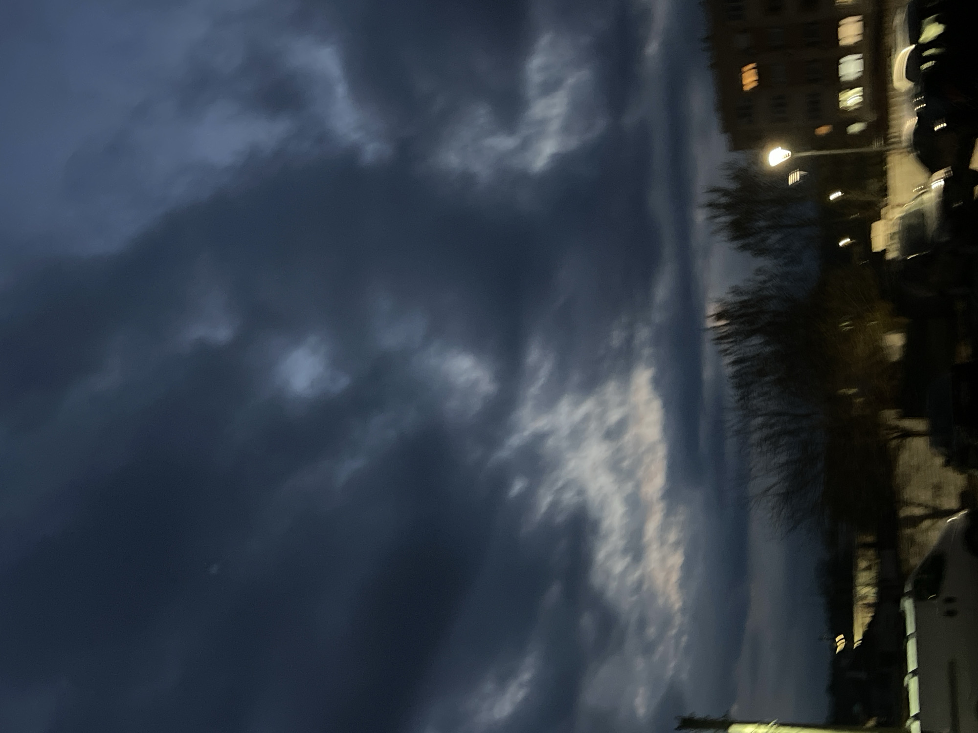 The image shows a nighttime scene in what appears to be an urban environment. On the right side, there are buildings with illuminated windows. A streetlight is visible, casting a glow on nearby trees and street-side objects, including cars and what appears to be a utility pole. The left side of the image is dominated by a dark, cloudy sky. The picture is taken in Elhovo, Bulgaria.
