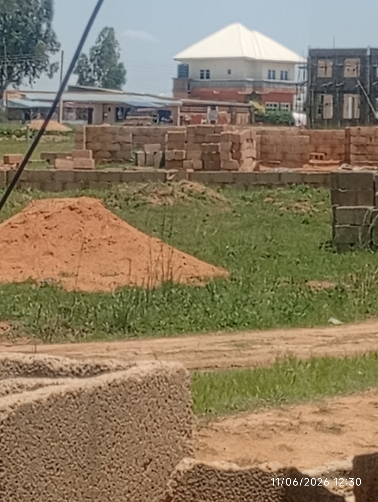 This image depicts a construction site in the city of DU, Nigeria, on June 11, 2026, at 12:30 PM. The foreground shows a pile of bricks and a mound of sand, suggesting ongoing construction work. In the middle ground, a partially constructed building with brick walls is visible. The background features a larger building with a white roof, possibly a residential house, and another building under construction with exposed windows and scaffolding. Trees and vegetation are present, indicating an outdoor setting. The sky is clear, suggesting fair weather. No people are visible in the image.
