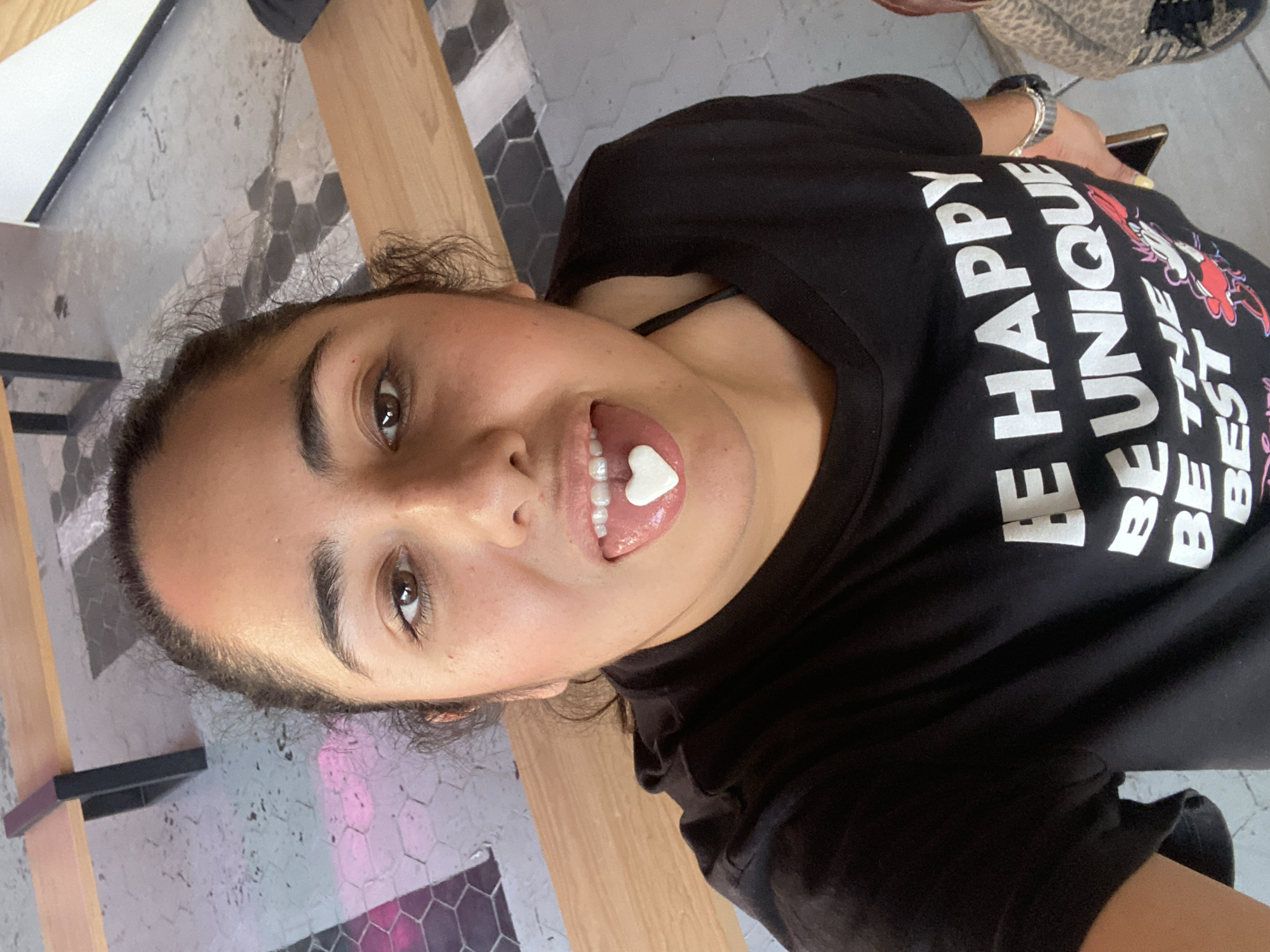A young person, likely female, is captured in a casual, rotated selfie or close-up shot, looking directly at the camera with a playful expression. She has dark hair and brown eyes, and is wearing a black t-shirt. On her extended tongue, a small, white, heart-shaped candy is visible.
Her black t-shirt features prominent white text, partially visible, that reads "BE HAPPY BE UNIQUE BE THE BEST," accompanied by a red and white cartoon character, possibly the Pink Panther. A watch is worn on her left wrist, and a smartphone is held in her left hand, partially obscured.
The setting appears to be an indoor or covered outdoor area, characteristic of a cafe or casual public space in Puebla, Mexico. The background includes light-colored wooden surfaces, possibly part of a bench or table. The floor is distinctive, covered with grey and black hexagonal tiles. A section of a white wall with a black border is also visible. In the upper background, a patterned object, possibly a leopard-print bag or shoe, can be seen. The lighting suggests it is daytime.