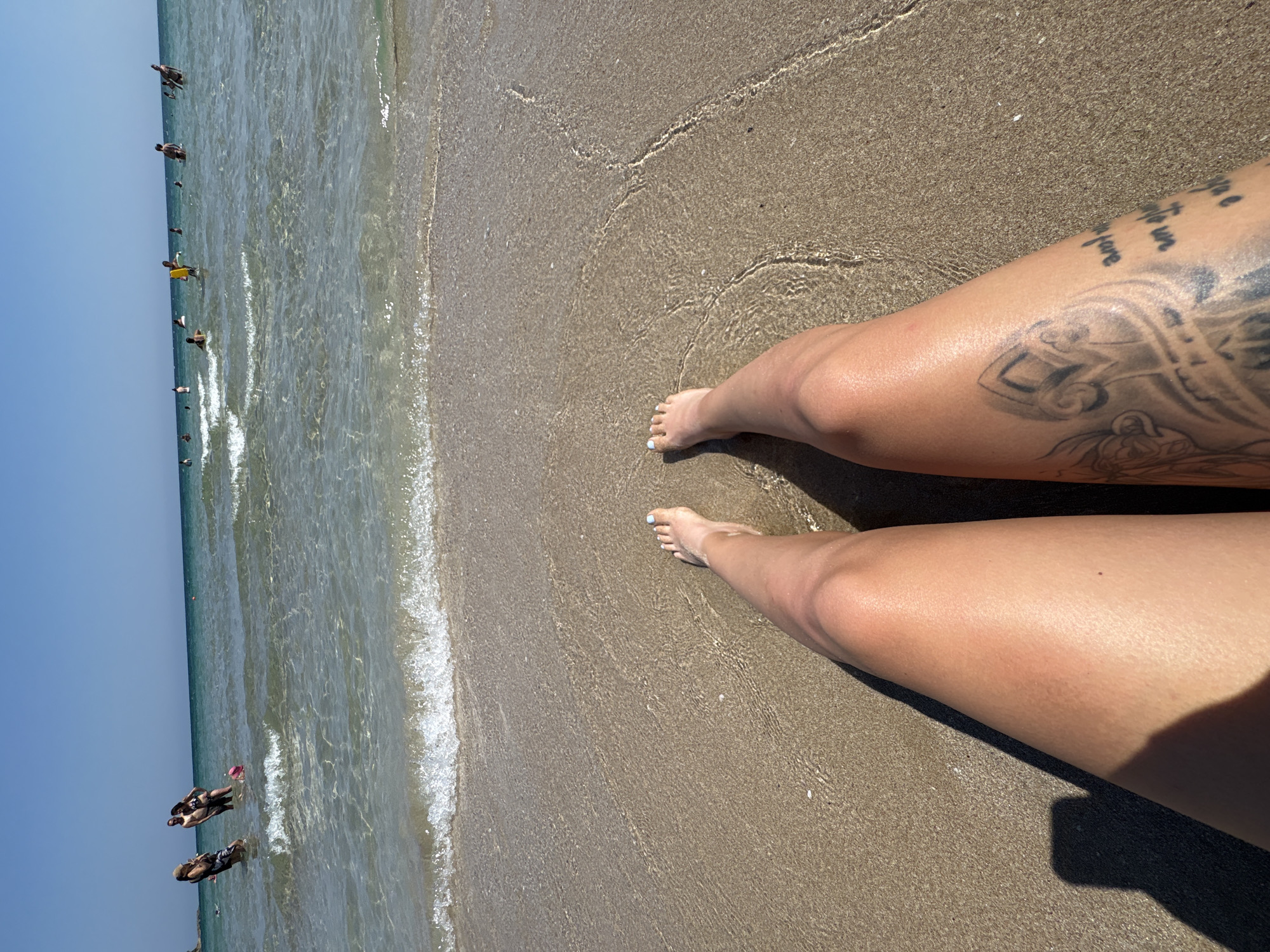 The image depicts a person's legs and feet on a sandy beach, with the shallow, clear ocean waves lapping at the shore. In the background, numerous people are visible swimming and wading in the sea under a clear blue sky. The location is identified as Primorsko, Bulgaria. The foreground is dominated by the sun-tanned legs of the person, one of which has a tattoo on the thigh. The sand is light brown and wet near the water's edge, showing ripples from the receding waves. The ocean water is transparent, allowing the sandy seabed to be seen. The overall scene conveys a summer day at the beach.