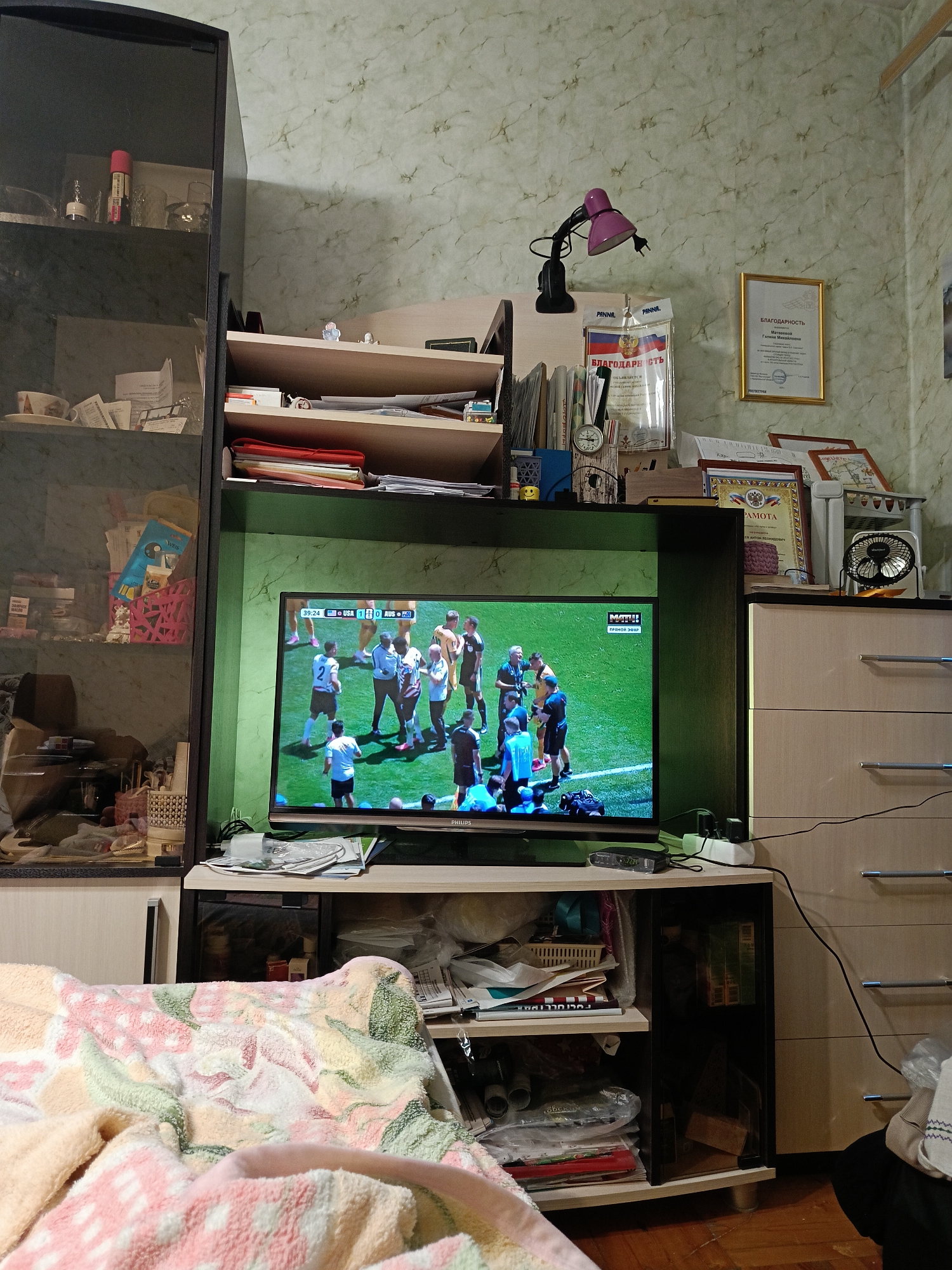 This is a full shot image of a living room with a television in the center displaying a soccer match. The room has a cluttered but lived-in feel. To the left, a tall, dark-colored cabinet with glass doors contains various items on its shelves, including what appear to be bottles, books, and decorative objects. To the right of the television, a dresser with light-colored drawers is visible. The walls are covered in a light-colored wallpaper with a subtle marbled pattern. Above the television, on a shelf, are several framed documents and awards, including a gold-framed certificate. A small pink desk lamp is also attached to this shelf. The television rests on a dark media console, which is also filled with papers and other miscellaneous items. In the foreground, a soft, patterned blanket is draped over what appears to be a sofa or chair. The lighting suggests it is either daytime or evening with indoor lights on. The presence of specific Russian writing on some of the documents and awards suggests a Russian context, potentially Arkhangelsk, Russia, based on the prompt.