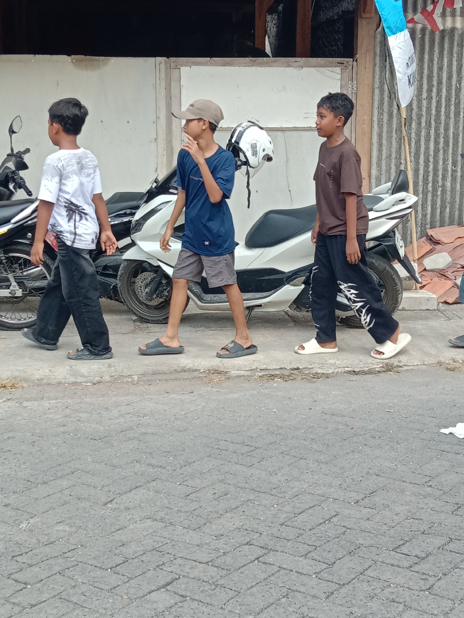 Three young boys are walking on a paved sidewalk in front of motorcycles and a building. The boy on the left wears a white t-shirt with a palm tree design and dark pants. The middle boy wears a blue t-shirt, grey shorts, and a beige baseball cap. The boy on the right wears a brown t-shirt and dark pants with white detailing. A white helmet is held by the middle boy. The building behind them is white with wooden trim and corrugated metal on the right side. A flag pole with a blue and white flag is visible. The ground is paved with grey interlocking bricks, and the area appears to be outdoors, possibly in a residential or commercial street in Wirosari, Indonesia.
