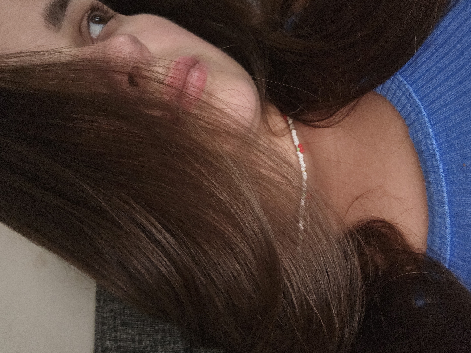 The image is a close-up, indoor portrait of a young woman. Her head is turned to the right, and her brown hair partially obscures her face. She is wearing a blue, ribbed top and a beaded necklace with small white, red, and green beads. The background consists of a textured grey surface and a lighter, off-white wall. The lighting appears to be artificial and soft. There are no observable elements within the image to suggest a specific location in the city of Petropavlivska Borshchahivka, Ukraine.