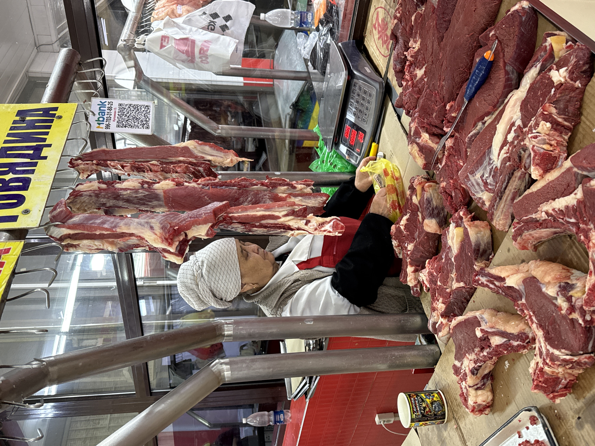 This image depicts a bustling meat stall or butcher shop in Bishkek City, Kyrgyzstan, likely during daylight hours. The central subject is a woman, presumably a butcher or vendor, captured from the waist up. She wears a white uniform, a grey sweater, and a white head covering, and is actively handling meat with a yellow plastic bag in her hands.
The stall is abundant with fresh, raw beef cuts, displayed both on a wooden counter and hanging from metal hooks. Adjacent to the meat is a digital weighing scale, its display showing numerical values (e.g., 0.000 and 3.5), along with a knife featuring a blue and orange handle.
Key textual details include a yellow sign with the Cyrillic word "ГОВЯДИНА" (GOVYADINA), translating to "Beef." Nearby, a white card presents a QR code for payment, identifying "mBank" and displaying a phone number "996-703-31-88-25" and the name "Шукурова О." (Shukurova O.). In the background, various plastic bags and bottles are visible behind a glass partition. A white cup adorned with "CHAMPION FALCON" text sits on a surface lower down.
The scene suggests a routine commercial transaction in a market setting, with the woman focused on her work. The overall atmosphere is functional and practical, characteristic of a fresh meat vendor.