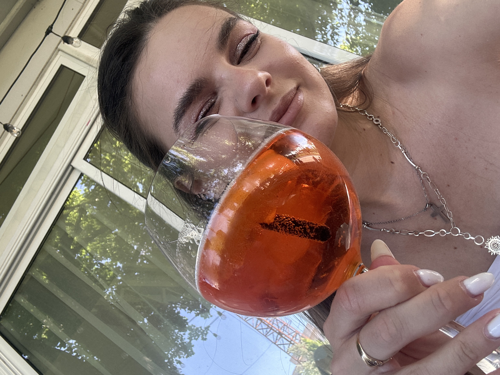 A woman is holding a large glass of orange liquid, possibly a cocktail, up to her face. She has her eyes closed and a slight smile. She has dark hair and is wearing silver eyeshadow and a layered silver necklace with a cross and a sunflower pendant. She is wearing a ring on her ring finger. Her fingernails are long and painted white. The background shows a window with sunlight streaming through, reflecting trees and what appears to be a construction crane in the distance. The scene is likely taking place in a restaurant or cafe in Plovdiv, Bulgaria based on the architectural style and the presence of the construction crane which can be associated with the reconstruction of the historical town in Plovdiv. There are string lights visible on the exterior of the building.