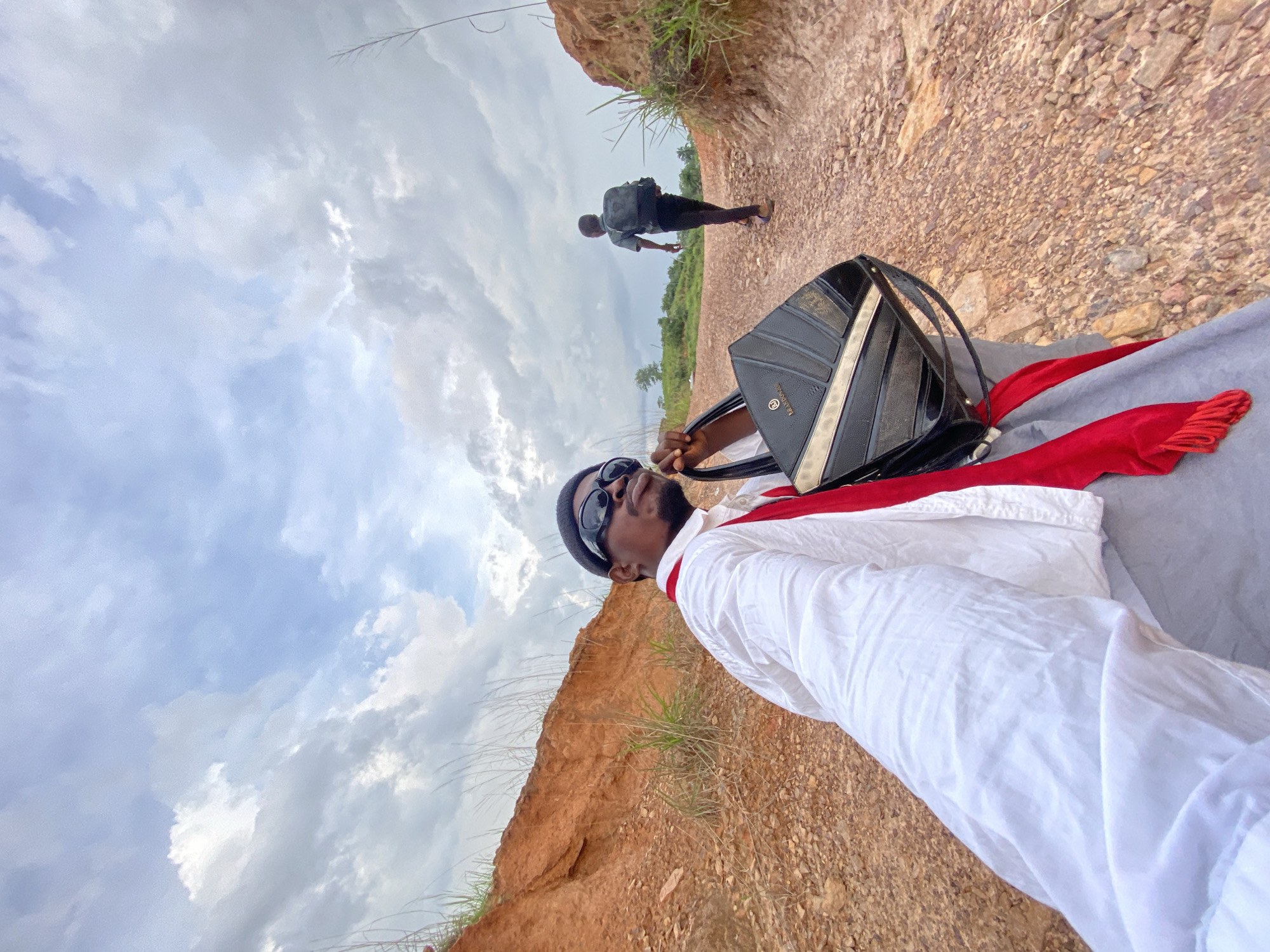 The image depicts an outdoor scene in Ufu Esioni, Nigeria, under a cloudy sky, captured with a tilted perspective. In the foreground and mid-ground, a person, likely male, is posing for the camera. They wear a white long-sleeved garment, a black beanie, and dark sunglasses, with a red fabric draped around their neck. Their left hand holds a black handbag adorned with vertical gold-colored stripes and some unidentifiable text, possibly a brand name, in gold lettering. The individual looks upwards and slightly to the right of the frame.
Further in the background, another person is walking away from the camera along a reddish-brown, gravelly path. This individual wears dark clothing and a backpack. The path is bordered by steep, eroded banks of reddish-brown earth with patches of green grass and some distant trees. The scene suggests an outdoor activity or journey through a natural, rugged landscape on an overcast day.