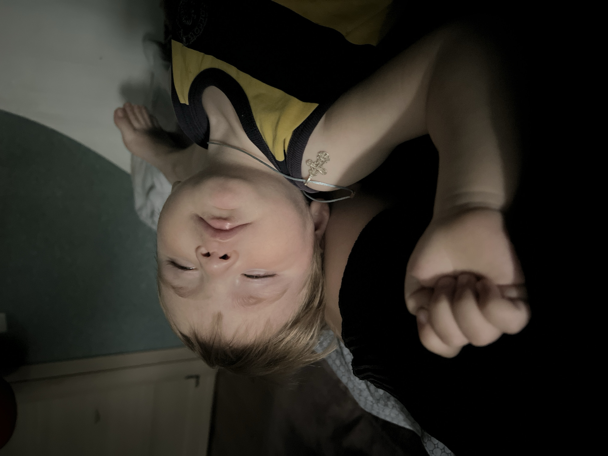 A young child with fair skin and blonde hair is lying on their back. The child is wearing a yellow and black shirt and a necklace with a cross pendant. The child's eyes are closed. The child's right hand is clenched into a fist. The child's feet are visible. The background includes a wall and a piece of furniture. There is not enough information to identify the specific location in Noviy, Russia.