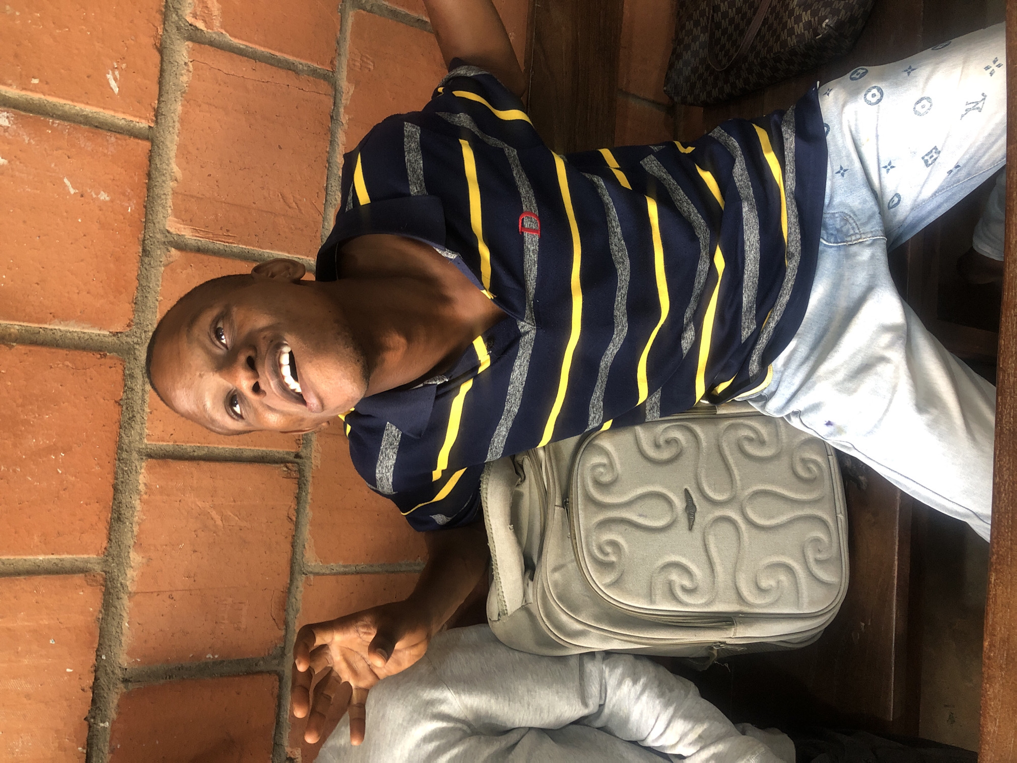An adult Black male sits on a wooden bench in front of a brick wall. He is wearing a navy blue polo shirt with horizontal yellow and grey stripes and light blue jeans with a pattern of repeating blue symbols. His left arm is outstretched with his hand open, and his right arm is bent upwards. He is looking up and to the right with his mouth open. A light grey backpack with a swirling pattern on it is on the bench to his left. To his right, a dark patterned bag rests on the bench. The background is an orange brick wall.