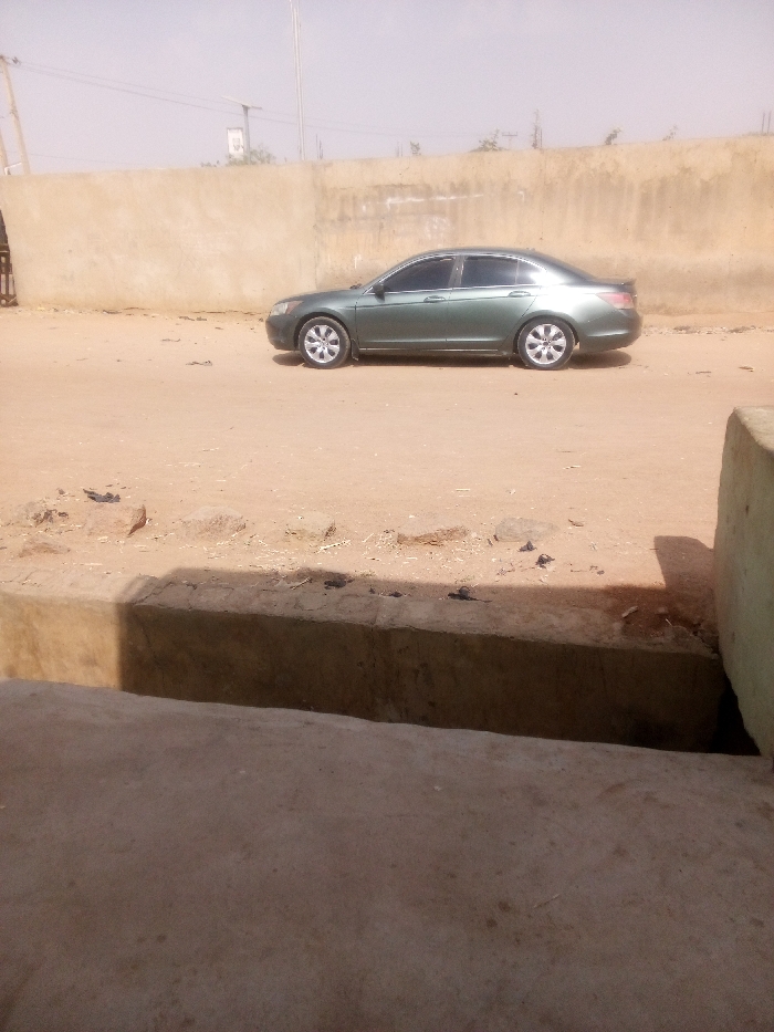 The image displays an outdoor scene in Tudun Murtala, Nigeria, under bright, hazy daylight. A light-green four-door sedan, consistent with an 8th-generation Honda Accord, is parked on an unpaved, light-brown dirt surface, oriented to the viewer's left. The vehicle has tinted windows, silver multi-spoke rims on all four wheels, and a black spoiler on the trunk lid. Behind the vehicle, a long, weathered light-brown wall extends horizontally across the midground. A dark, possibly metal, gate structure is partially visible on the far left, integrated into the wall. The immediate foreground features a concrete surface, which casts a shadow, and an adjoining concrete drainage channel or curb that separates the foreground from the dirt road. Small rocks and debris are scattered across the dirt ground. The distant background reveals a hazy sky with faint outlines of utility poles and lines. No individuals are present in the scene.