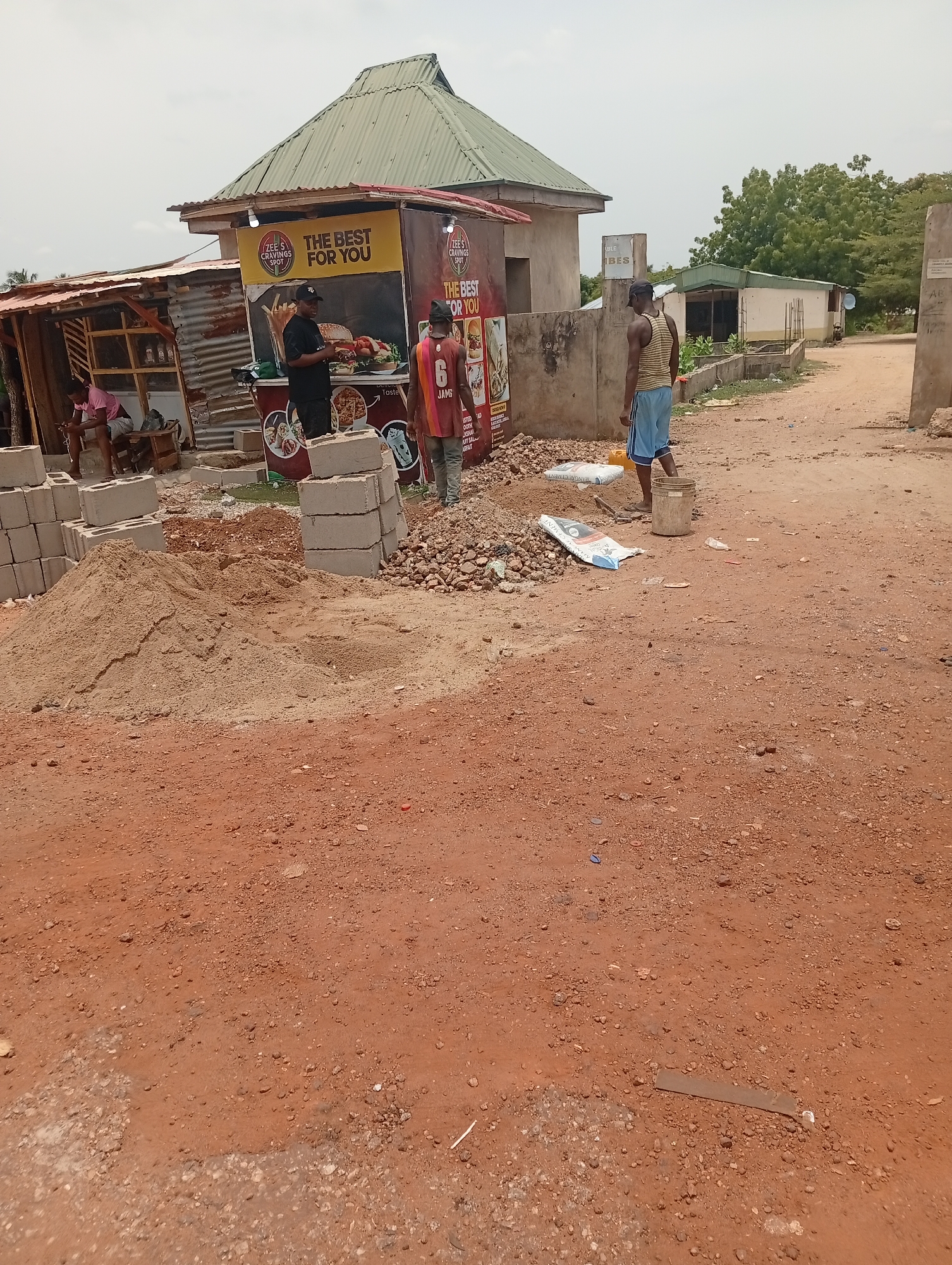 The scene captures an outdoor construction site in Adankolo, Nigeria. The ground is a reddish-brown, dirt-covered area with scattered small stones. In the mid-ground, there is a pile of sand and several stacks of grey concrete blocks. A ripped bag, possibly cement, lies on the ground amidst a pile of gravel and rubble. Three men are visible in the scene. One man is standing near a food stand. Another man is wearing a striped tank top and blue shorts, carrying a bucket. He is located on the right side of the frame and is walking towards a building in the background. A man in a red jersey is standing on the left, near some cinder blocks. To the left is a shack-like structure made of corrugated metal, where a person is sitting. Behind these figures is a food stall with the signage "ZEE'S CRAVINGS SPOT: THE BEST FOR YOU" on its front. Other buildings and greenery are visible in the background. The sky appears overcast.