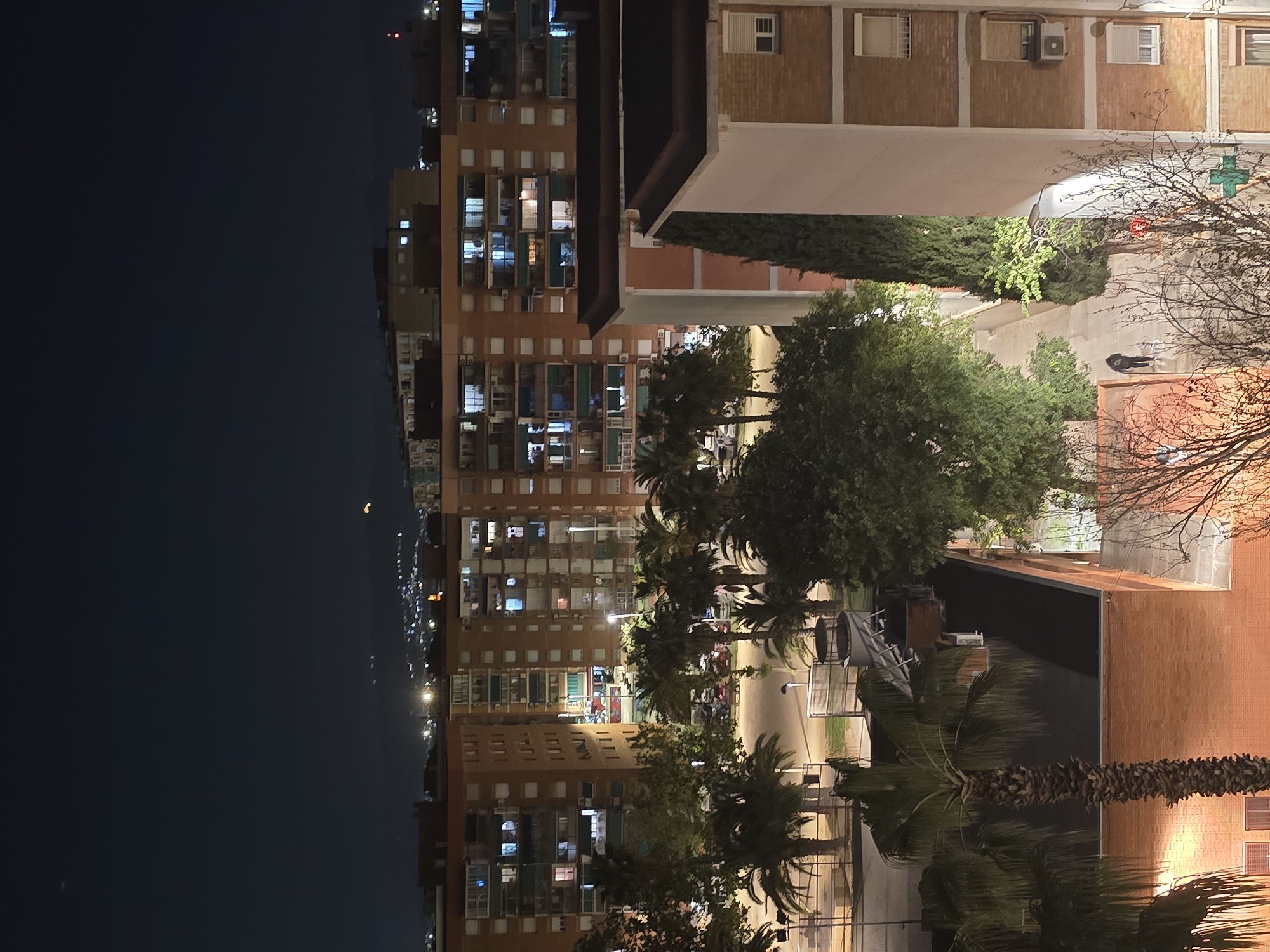 The image shows an urban scene at night in Cornellà de Llobregat, Spain. Several tall apartment buildings with illuminated windows dominate the left and center of the frame. The sky is dark. A street lined with palm trees and other trees runs through the center. On the right, there are brick buildings with visible air conditioning units and windows. A courtyard or park area with trees, a path, and possibly a person walking is present between the buildings. The perspective is slightly elevated, as if taken from a higher floor of a building or from a raised platform.