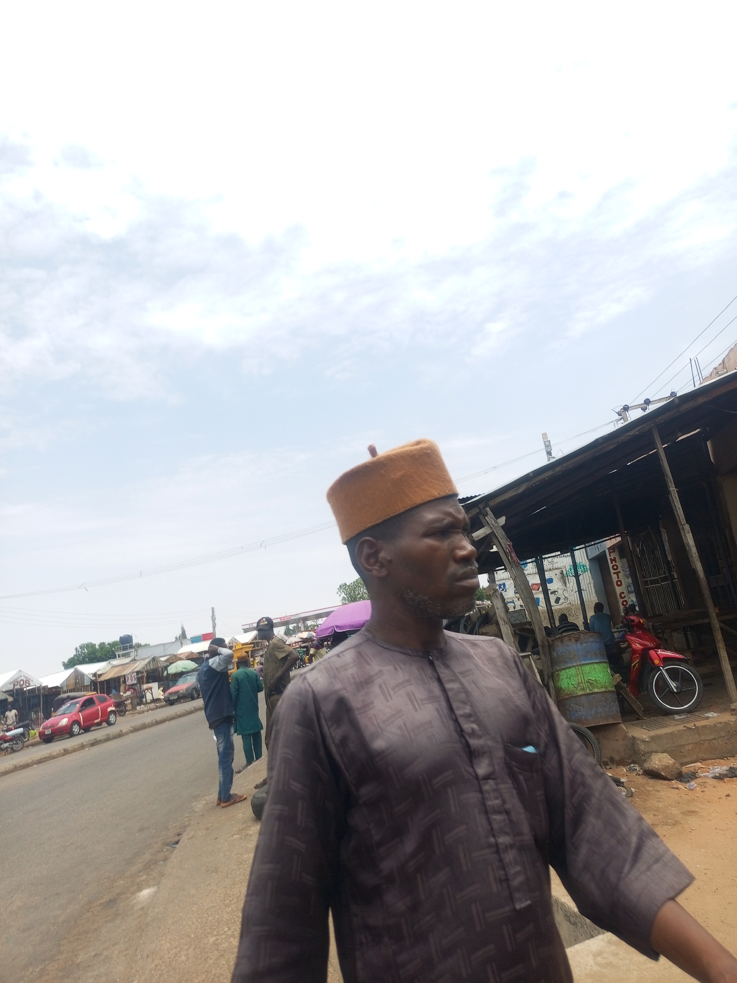 The image depicts a man in Gombe, Nigeria, dressed in a brown traditional shirt and a brown cap. He stands near a roadside with buildings and shops in the background. A red car is visible on the street. Other people are present in the vicinity. A red motorcycle is visible near a dilapidated structure. The sky is overcast.