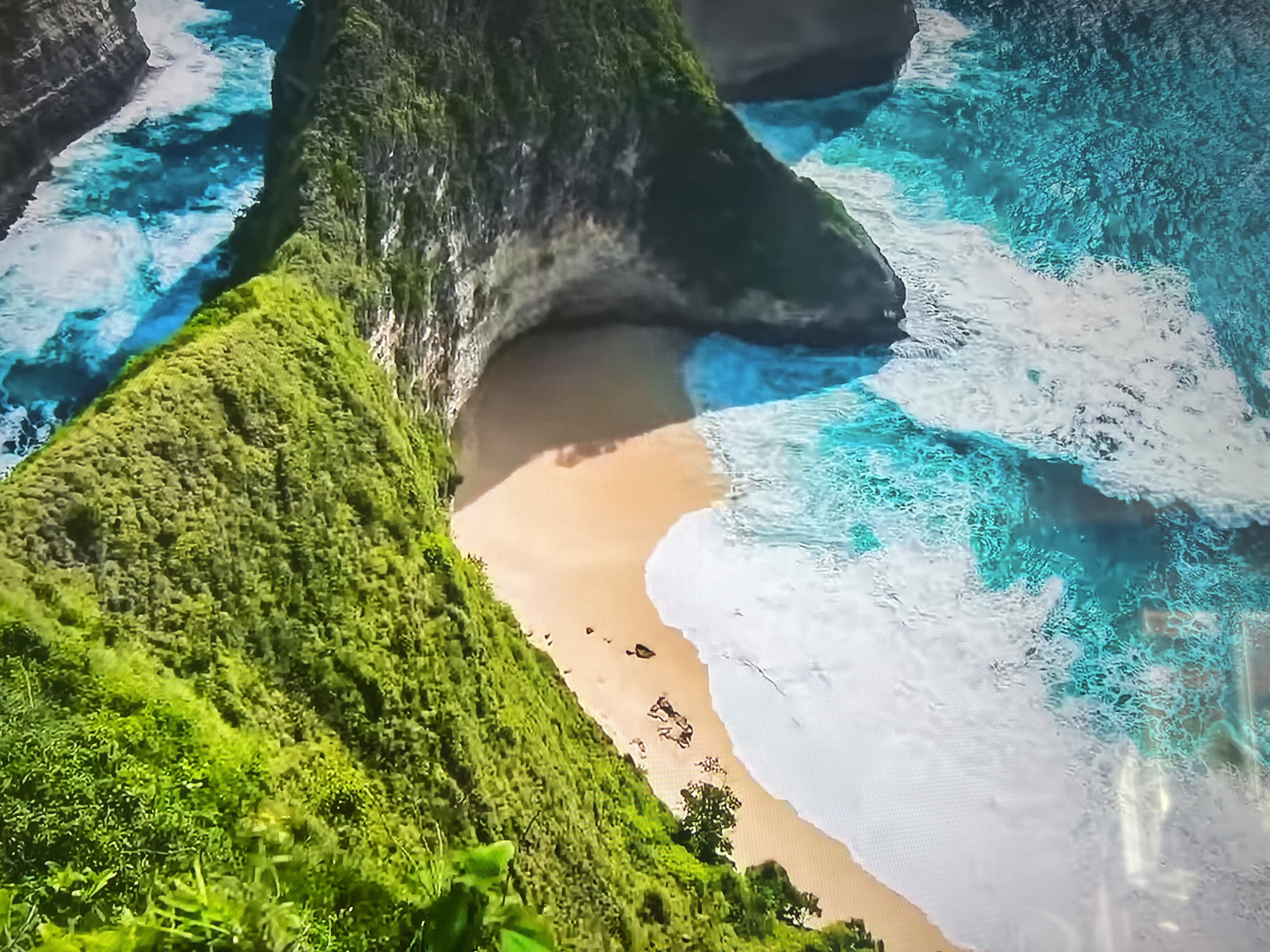 The image shows an aerial view of a tropical beach with turquoise water and white waves crashing against a sandy shore. Lush green vegetation covers the cliffs surrounding the beach. There are no indications that this image is in Basingstoke and Deane, United Kingdom.