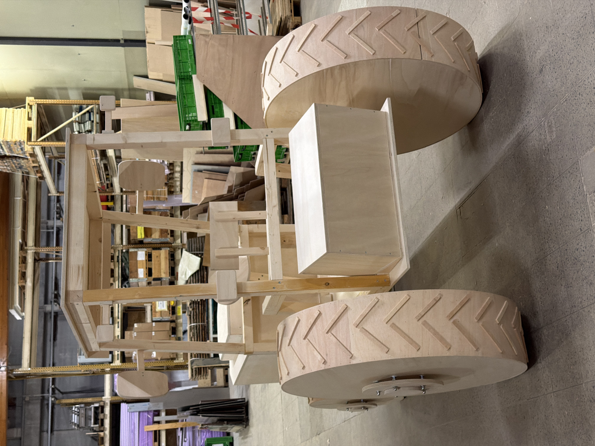 The image shows a wooden model of a tractor in a workshop. The tractor is unfinished, with exposed wooden planks and supports. It has large, ridged wheels that mimic tire treads. The background reveals a warehouse environment with shelves stacked with materials, including crates and wooden panels. There are no people visible in the image. The location is specified as a workshop in Leudelange, Luxembourg.