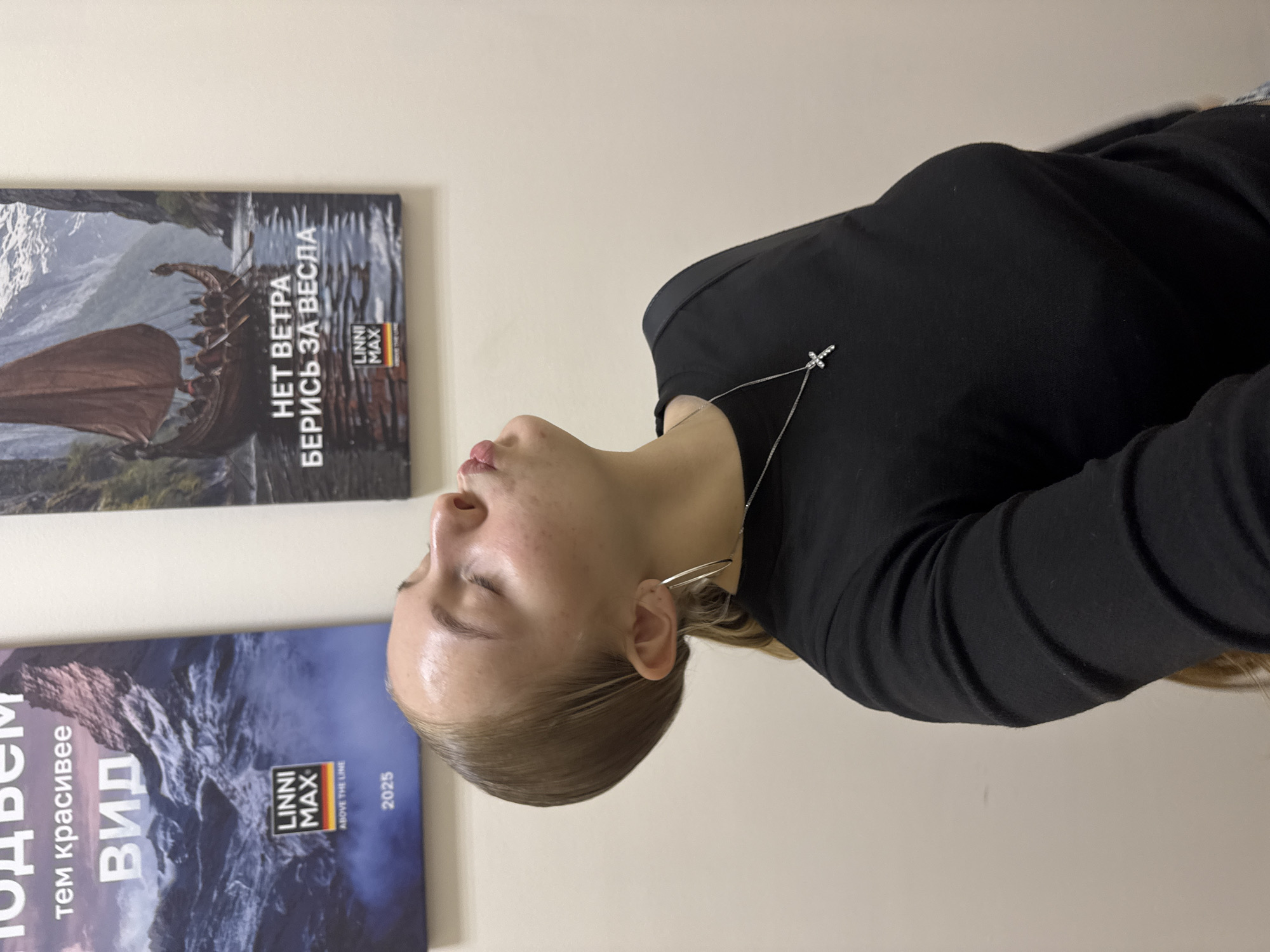 A young woman with blonde hair pulled back into a ponytail and wearing a black long-sleeved shirt and a silver cross necklace, poses for a selfie in front of two posters on a beige wall. The poster on the left features a mountainous landscape with the Russian text "тем красивее ВИД" and a logo that reads "LINNI MAX ABOVE THE LINE 2025". The poster on the right shows a Viking ship sailing on water with the Russian text "НЕТ ВЕТРА БЕРИСЬ ЗА ВЕСЛА" and a similar "LINNI MAX" logo. The lighting appears to be artificial, suggesting an indoor environment, possibly a gallery or exhibition space. No specific location in Perm, Russia, can be identified from the visual information provided.