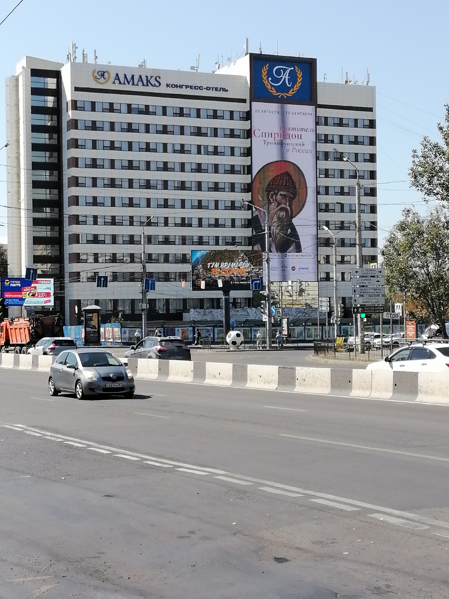 The image shows a wide street in Rostov-on-Don, Russia, with a large building complex, the congress hotel "AMAKS", dominating the background. The hotel has a distinctive sign with a wreath and the letter "A". A large banner on the building advertises an exhibition titled "Святитель Спиридон. Триумф в России" (Saint Spiridon. Triumph in Russia) running from August 20th to September 20th. Several cars are visible on the street, including a gray Toyota Yaris in the foreground. Traffic lights and road signs are present, indicating traffic flow and directional information. A large inflatable soccer ball is placed on the left side of the street, near a billboard. The sky is clear and blue, suggesting a sunny day. The scene depicts a typical urban environment with commercial and residential infrastructure.