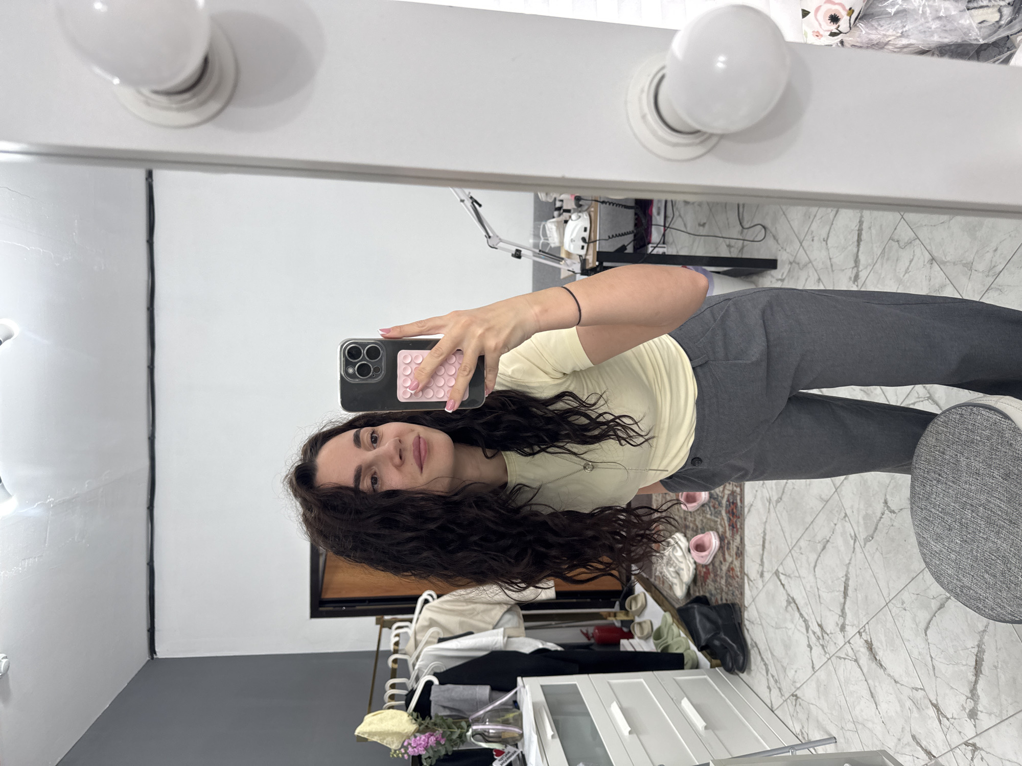 A young woman is captured in a mirror selfie, standing upright and looking directly at the camera with a slight smile. She has long, dark, wavy hair and is wearing a light yellow short-sleeved top and dark grey trousers. Her fingernails are painted light pink. She holds a smartphone with a dark case and a light pink textured accessory on its back.
The setting appears to be an indoor room, likely a bedroom or dressing area, within what is given as Chaikovsky, Russia. Behind her, reflected in the mirror, is a vanity mirror frame with two prominent white light bulbs. The room features a light-colored, marble-patterned tiled floor where various shoes are visible, including pink slippers, white sneakers, and black boots, alongside a small patterned rug. To the right, a gray upholstered stool or chair is partially visible.
In the background reflection, there's a white dresser or cabinet with clothes on hangers, a vase with flowers, and other items on top. A black cable runs down a corner of a white wall. The overall impression is that of a casual moment captured in a personal, well-lit space. No discernible text is visible in the image.