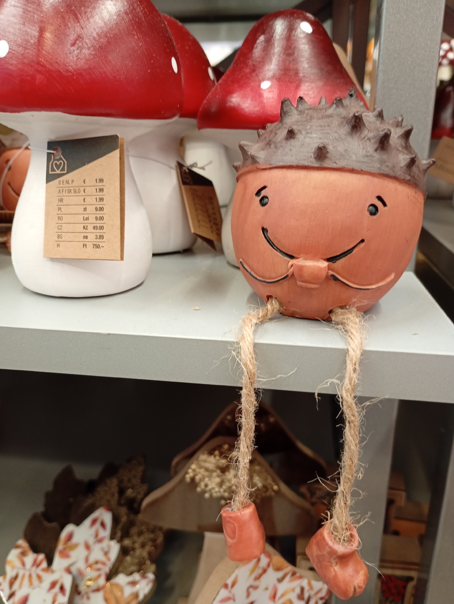 In a retail environment within Bad Lauterberg im Harz, Germany, a close-up shot focuses on decorative items displayed on a grey shelf. To the left, a large, white ceramic mushroom with a red cap, adorned with white polka dots, is visible. Attached to its stem is a price tag displaying various currencies and prices, including "DENLP € 1.99", "HR € 1.99", "PL zł 9.00", "RO Lei 9.00", "CZ Kč 49.00", and "H Ft 750.-". To the right of the mushroom is a small, terracotta-colored decorative figurine with a spiky brown head resembling a chestnut shell. The figurine has a simple, smiling face with black dots for eyes and a curved line for a mouth, and it is depicted with two dangling legs made of rough twine, each ending in a small, orange boot-like accessory. The background is blurred but suggests other shelves with more merchandise, including what appears to be autumnal-themed decorative leaves on the lower shelf. The lighting is diffused, creating soft shadows and highlighting the textures of the objects.