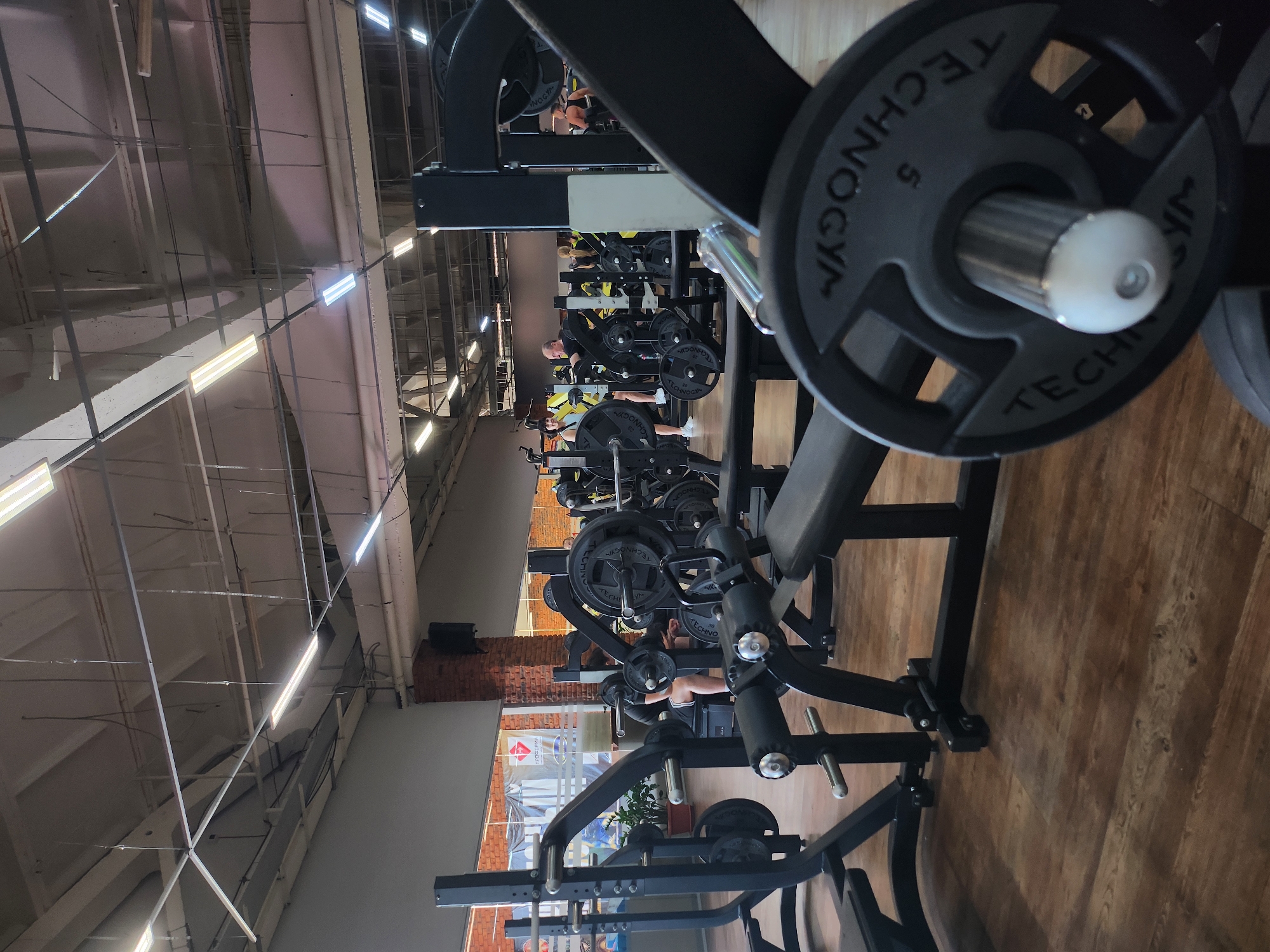 The image captures an indoor scene in a gym, featuring numerous weight-lifting machines arranged in a row. People are using the machines, focusing on their workouts. The equipment, primarily black in color, contrasts with the light-colored walls and ceiling of the gym. The flooring is wooden, and the gym has large windows that allow natural light to come in. The scene suggests a typical day at a well-equipped fitness center.