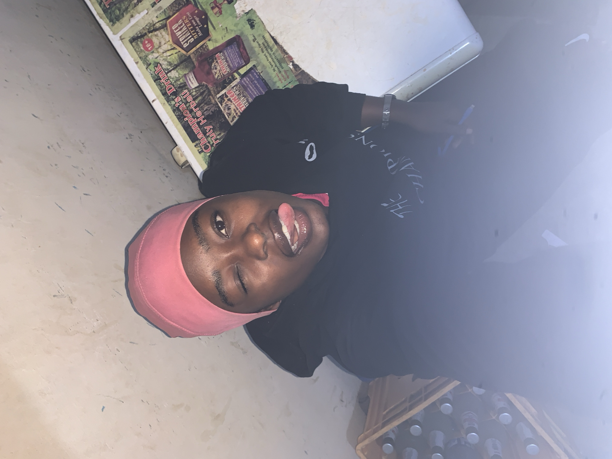 The image shows a person with dark skin wearing a pink head covering and a black shirt. The person is sticking their tongue out and winking. The background includes a wall, a cooler or refrigerator, a poster with the words "Champion's Drink Truly Herball STRIKER BITTERS" and other text, and a crate filled with bottles. The poster references the location as 50 Iyesi Road, Ota Ogun State, Nigeria.