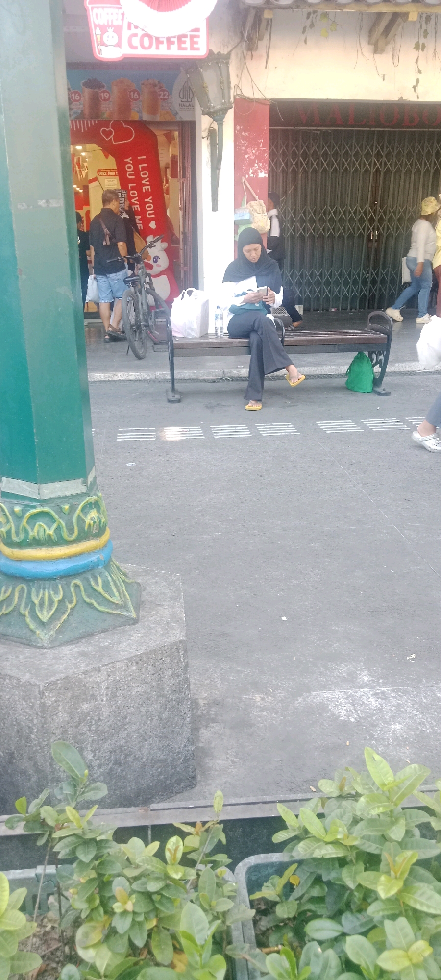 The image captures a street scene in Yogyakarta, Indonesia, likely near a commercial area. A green pillar with decorative accents stands on the left foreground, partially obscuring a shop entrance. The shop features a red archway with "I LOVE YOU" text and displays beverages. A man in a black shirt and denim shorts stands near the entrance, and a bicycle is parked beside him. To the right, a woman wearing a hijab and dark pants sits on a bench, looking at her phone. She is wearing yellow sandals. Behind her, a green bag rests on the ground. The background shows a building with a metal grate and more people walking by. In the lower foreground, potted green plants are visible. The ground is paved, with white tactile paving lines indicating a pedestrian pathway.