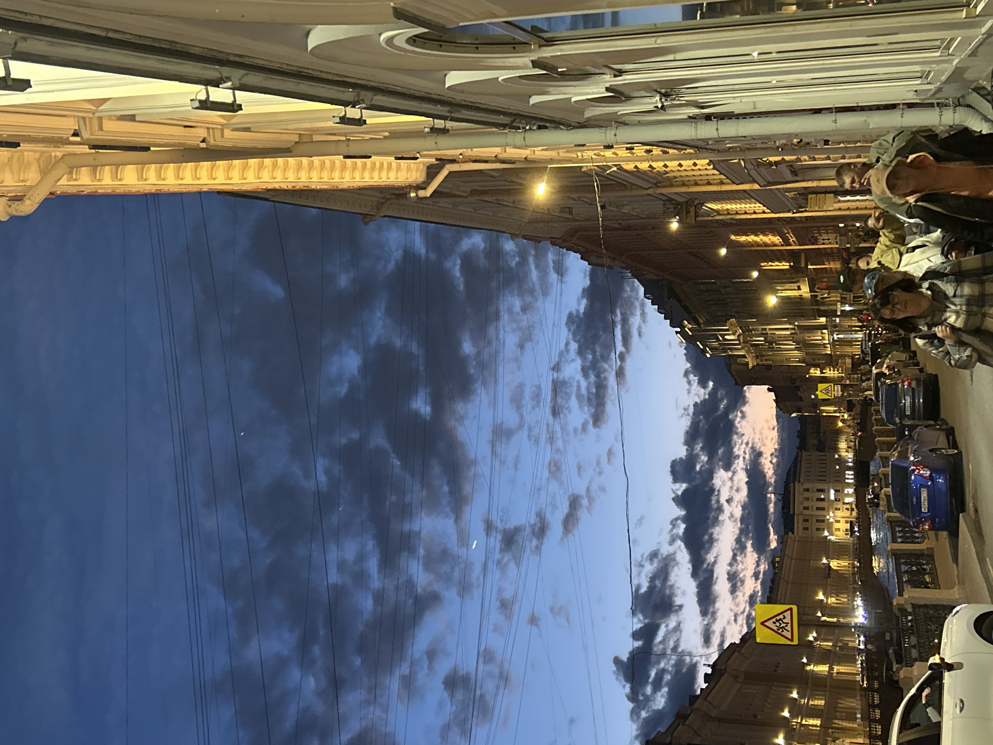 In the city of Unknown, Unknown, an urban scene unfolds at dusk along a canal. The sky is a deep twilight blue, transitioning to hues of orange and pink near the horizon, dotted with scattered clouds and crisscrossed by power lines. Along the canal, buildings with illuminated windows line the streets, casting a warm glow. A bridge spans the canal in the background, with more buildings and streetlights visible beyond.
On the right side of the image, a pedestrian walks along the sidewalk, facing away from the camera. Their clothing is not clearly discernible. Further down the street, a group of people are walking, also facing away from the camera. One individual, wearing a dark jacket and a light-colored baseball cap, is visible from the side. Another person in the group wears a plaid shirt and glasses.
On the left side of the image, cars are parked along the street, including a white car in the foreground. The street is lined with a decorative metal railing separating the sidewalk from the canal. Traffic signs are visible, including a yellow triangular sign with a pictogram of two people running. The overall atmosphere suggests a lively urban evening, with the city illuminated against the fading daylight.