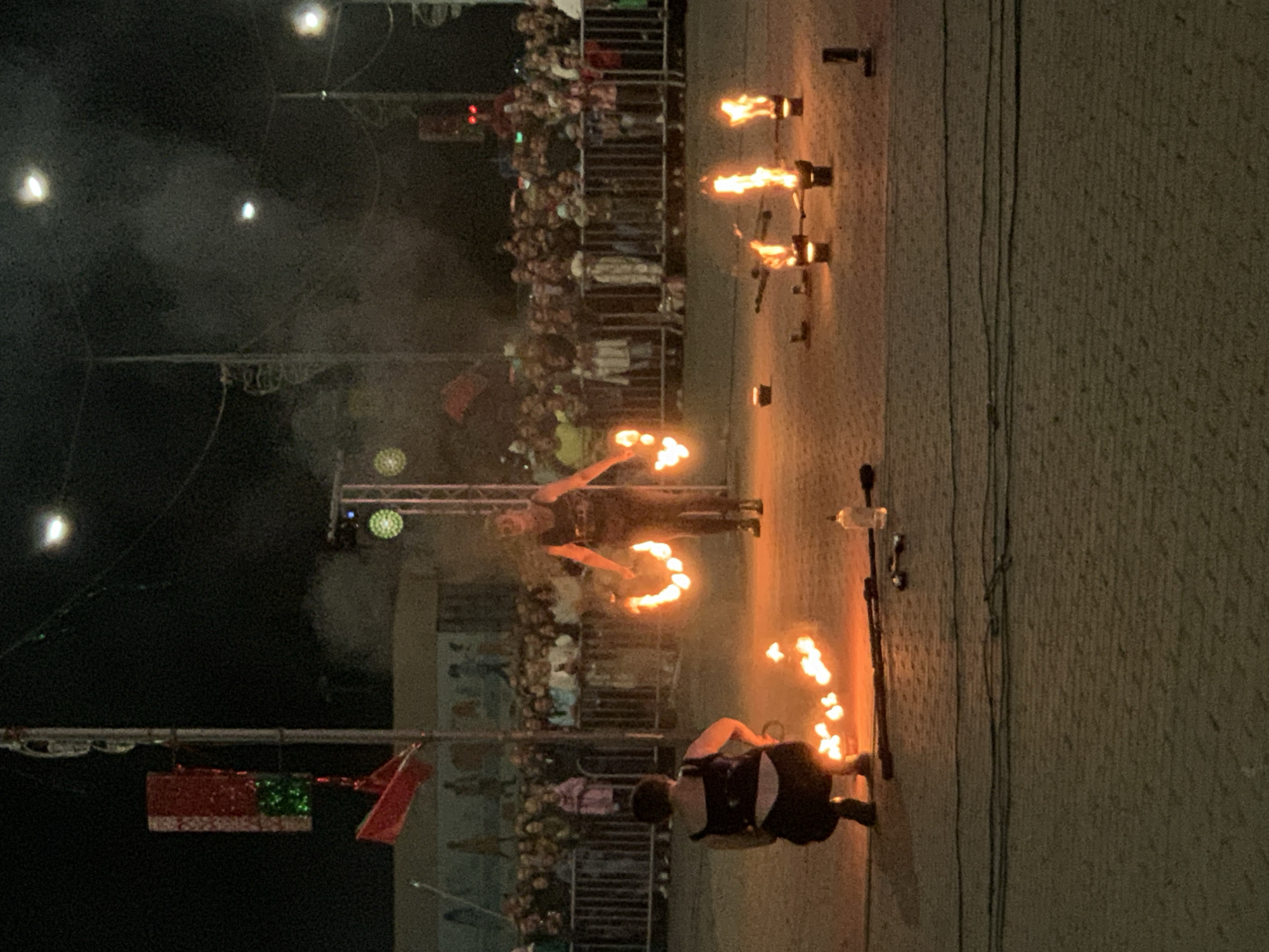 The image depicts a nighttime outdoor fire performance in the city of Kapyĺ, Belarus, likely near the central area given the crowd and urban elements. A performer, dressed in dark clothing, stands center stage, holding flaming props in each hand. They appear to be twirling the props, creating circles of fire. To the left, a second performer is crouched down, also interacting with flames on the ground, possibly preparing a routine or cleaning up. Several other flaming objects are positioned on the ground in front of the performers, adding to the spectacle.
A dense crowd of spectators is visible in the background, separated from the performance area by a metal barrier. The crowd is illuminated by stage lighting and the glow of the fire. Above the crowd, various elements of urban infrastructure are present, including poles, wires, and what appear to be decorative lights or signage. The overall environment suggests a festive or public event, such as a festival or street performance. The ground is paved, and the lighting is low, typical of an evening event. Smoke is visible in the air, likely a byproduct of the fire performance.