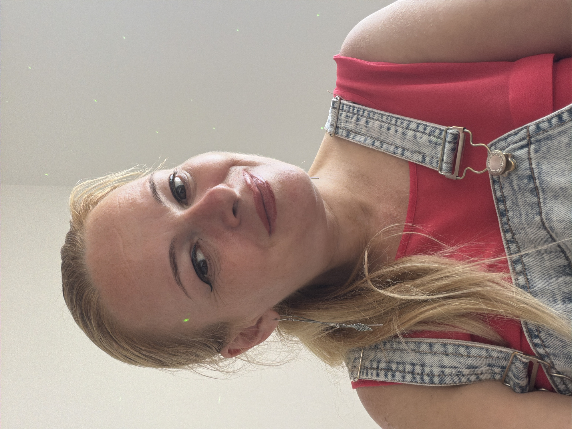 A medium close-up shot shows a white woman with blonde hair pulled back in a ponytail. She is wearing denim overalls over a bright pink shirt. The woman is looking towards the camera with a slight smile. There are several small green lights scattered in the background, which appears to be a plain wall.