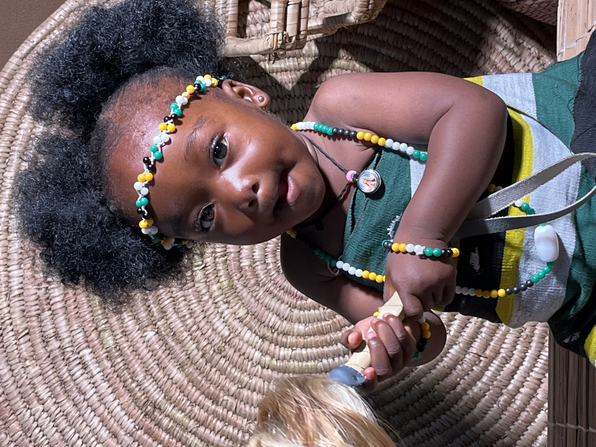A young Black child with dark hair and a beaded headband is lying on a woven mat. The child is wearing beaded necklaces and bracelets of varying colors. They are wearing a green, yellow, white, and black striped garment. The child is holding a brush with blonde hair. The location is unknown. There is no indication of the city of Barakin Akawo, Nigeria.