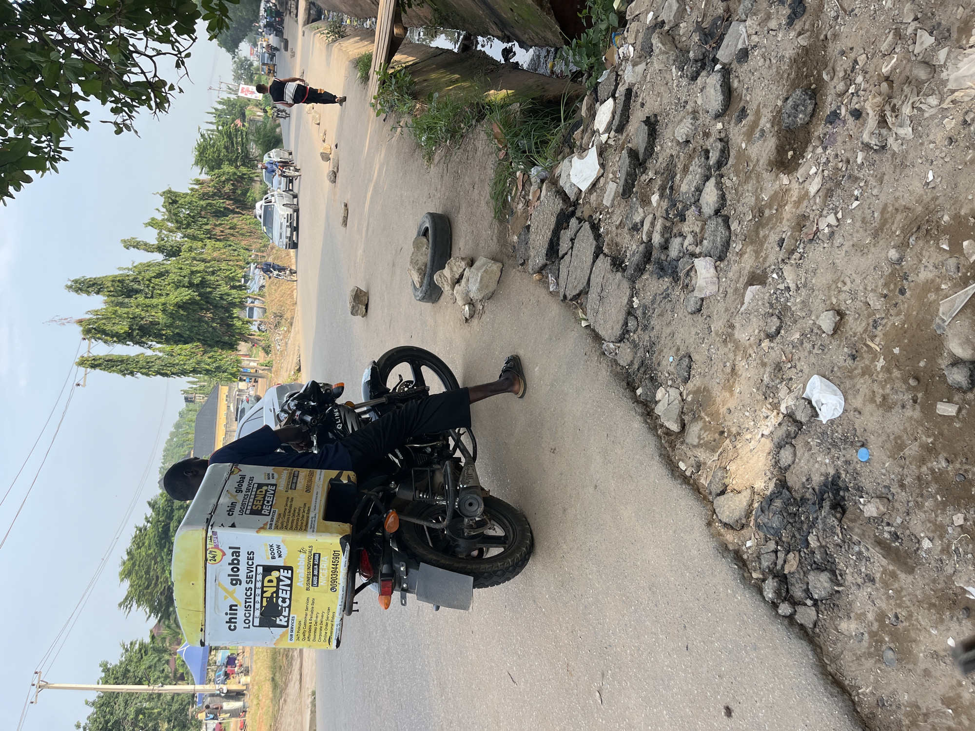 The image shows a street scene in what appears to be a city or town. A man on a motorcycle is situated on the right side of the road. The motorcycle has a delivery box attached to the back, labeled "chin global LOGISTICS SEVICES EXPRESS SEND RECEIVE". Text on the box includes "OUR SERVICES" and "Available for BUSINESS INDIVIDUALS LAGOS-ABUJA-ASABA NIGERIA," with the phone number 09039445901.
The road is paved, with visible cracks and damage along the edges, where there is an area of dirt, debris, and broken pavement. An old tire and several rocks are in the middle of the road. A drainage ditch with some water is located next to the road.
In the background, there are parked cars and trees. A person is walking on the road. The scene is set in daylight under a somewhat hazy sky.