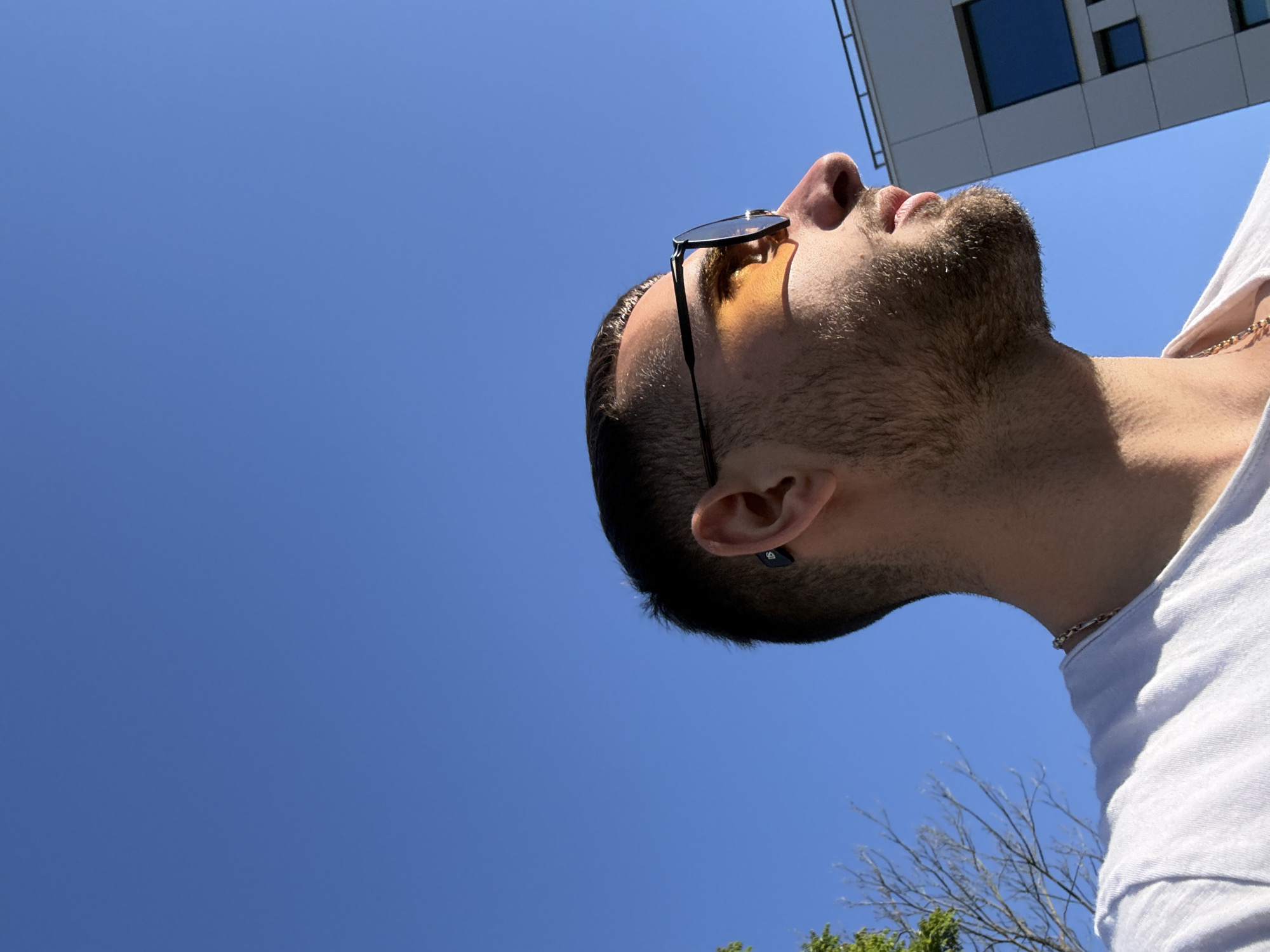A low-angle shot captures a man with tan skin, a trimmed beard, and short dark hair, wearing sunglasses with yellow lenses and a silver chain necklace. He is gazing upwards with a building partially visible at the top-right corner. He is wearing a white t-shirt. A small portion of tree branches and green foliage are visible in the lower right corner. The background is a clear, bright blue sky. The location appears to be urban, potentially in Gabrovo, Bulgaria.