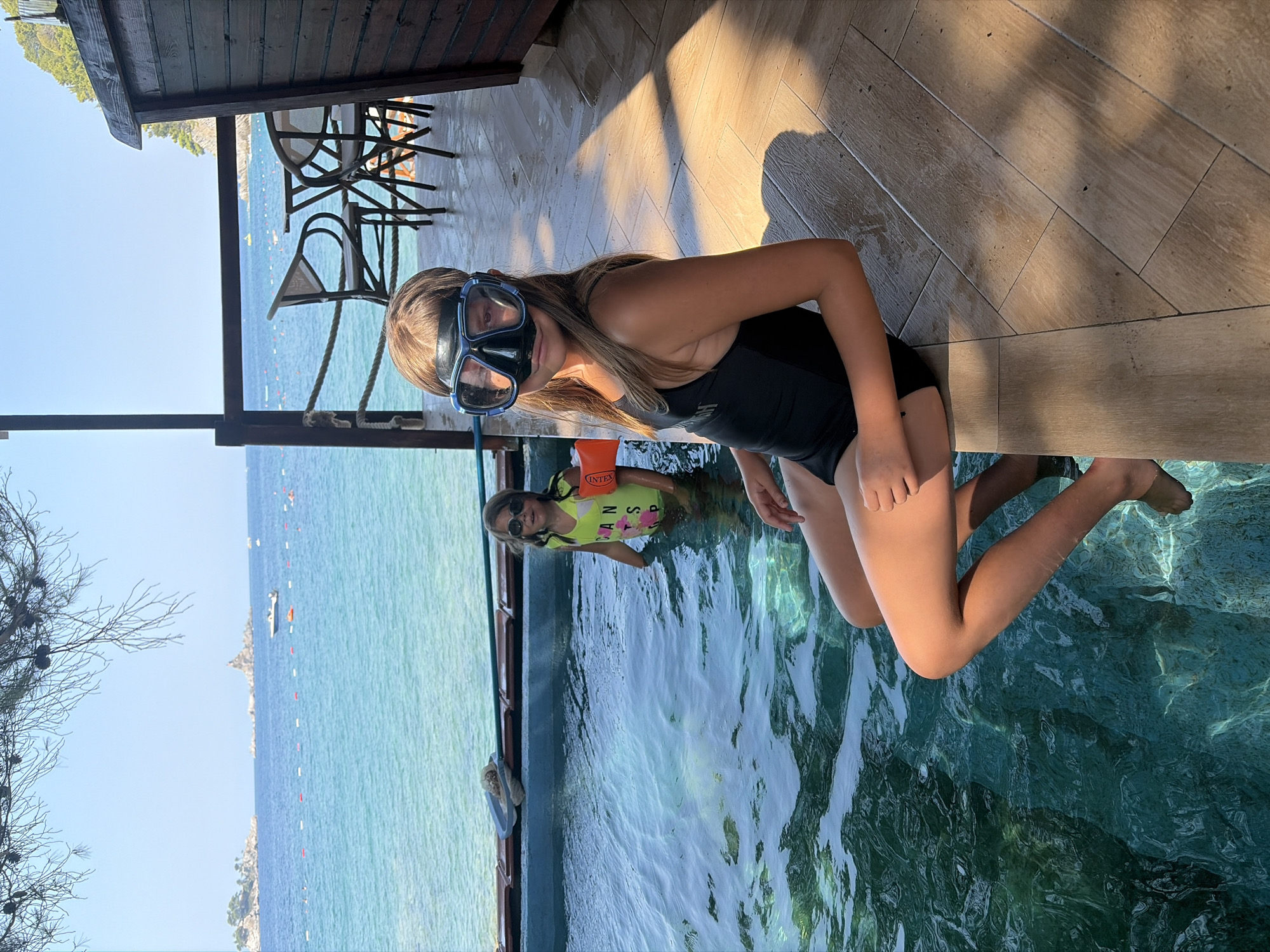 The image shows two young girls in a shallow body of water, likely a pool or a very calm sea area, with a wooden deck and railings in the foreground. The girl closer to the camera is wearing a black swimsuit and a diving mask. Her arms are resting on her knees as she sits on the edge of the water. The other girl is in the water, wearing a bright yellow life vest with "INTEX" printed on it and sunglasses. She has on a light-colored t-shirt with a floral print and some visible text that appears to read "CAN'T STOP".
The setting is outdoors, with the sea and a distant, hilly coastline visible in the background. The water is clear and inviting. Sunlight is casting strong shadows on the wooden deck, suggesting it is daytime, likely mid-morning or afternoon. The overall mood appears relaxed and playful, with the girls ready to enjoy aquatic activities. The presence of beach chairs under a wooden structure hints at a resort or a waterfront establishment. The scene is set in Petrovac centar, Montenegro.