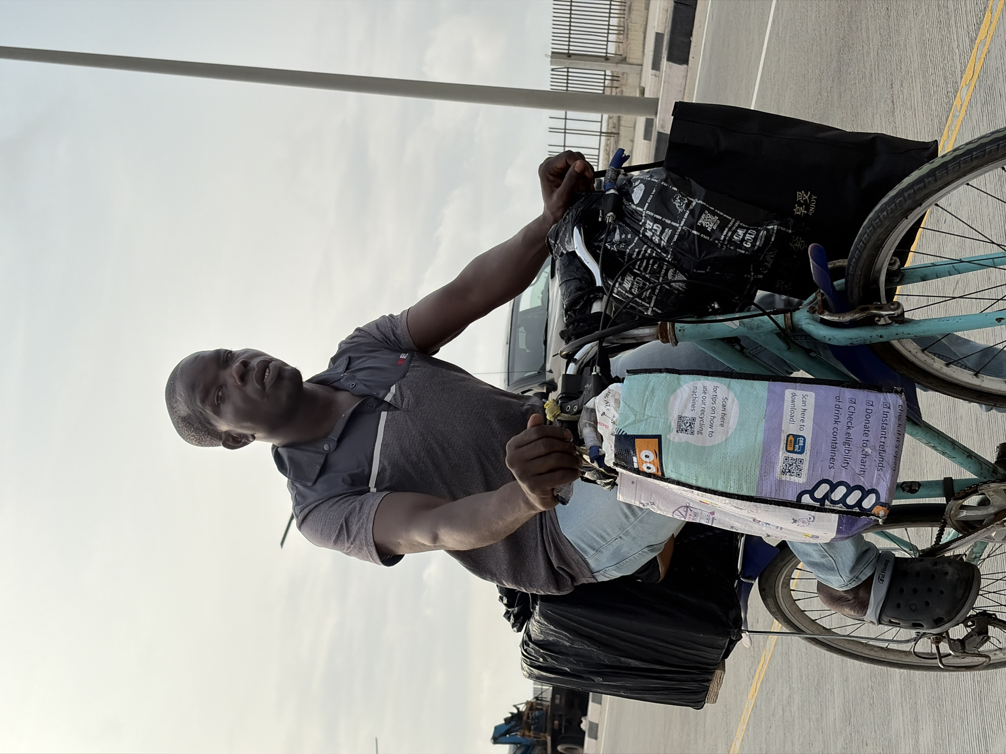 A Black man is riding a bicycle laden with large black plastic bags and other items strapped to the rear rack. He is wearing a grey and black polo shirt and light blue jeans, and black Croc-style shoes. The bicycle is turquoise. In the foreground, a colorful reusable bag with text and graphics is attached to the handlebars. In the background, a construction vehicle is visible, as well as a fence and a light pole. The scene appears to be on a paved road with yellow lane markings. The sky is overcast. The location is Itirin, Nigeria.