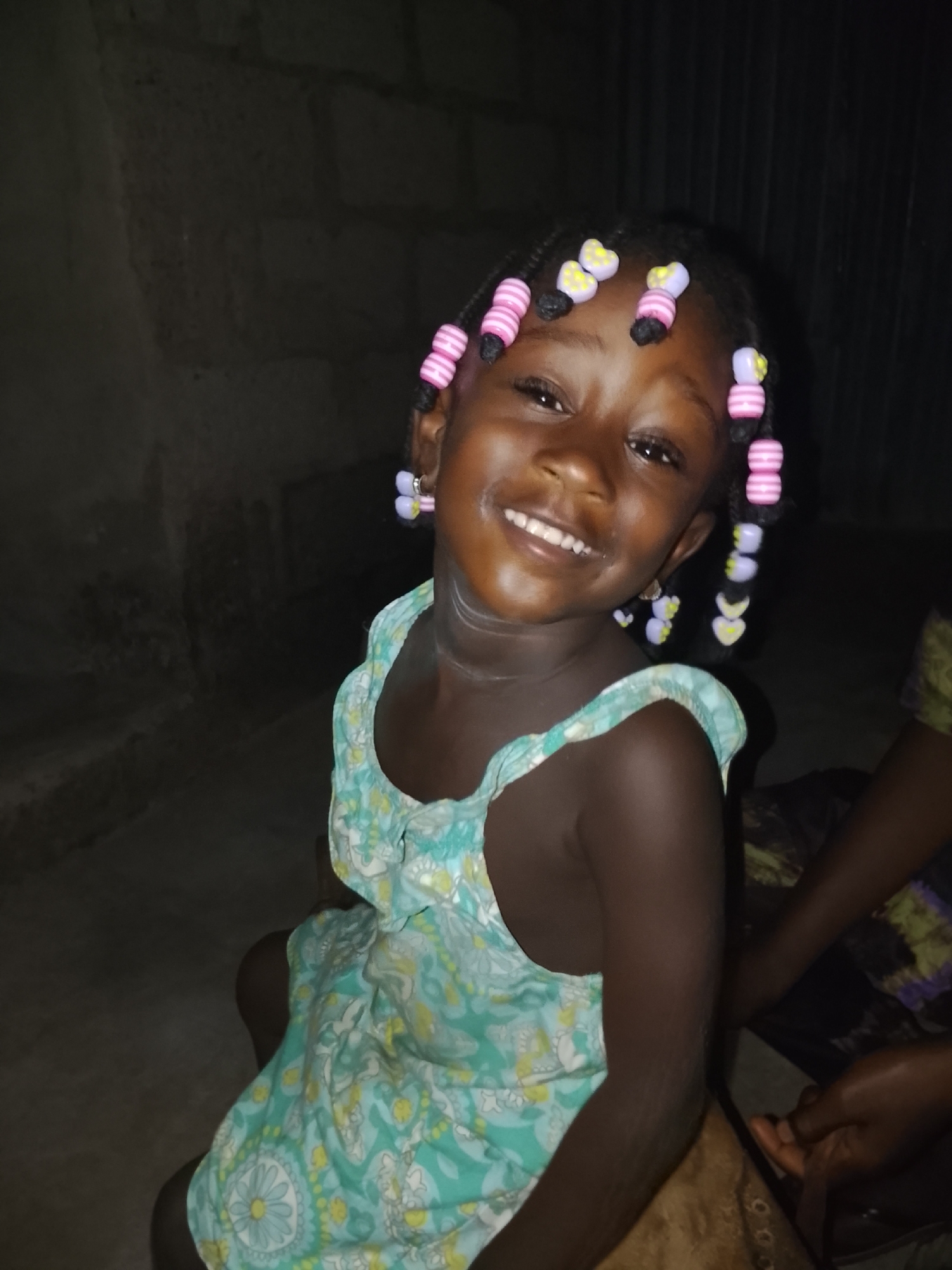 A young Black girl is sitting in an indoor location. She is wearing a light blue and white dress with a floral pattern. Her hair is braided and adorned with pink and yellow beads. The background appears dark and indistinct, but includes a dark wall to the left and a second person partly visible to the right. The girl is smiling and looking towards the camera.