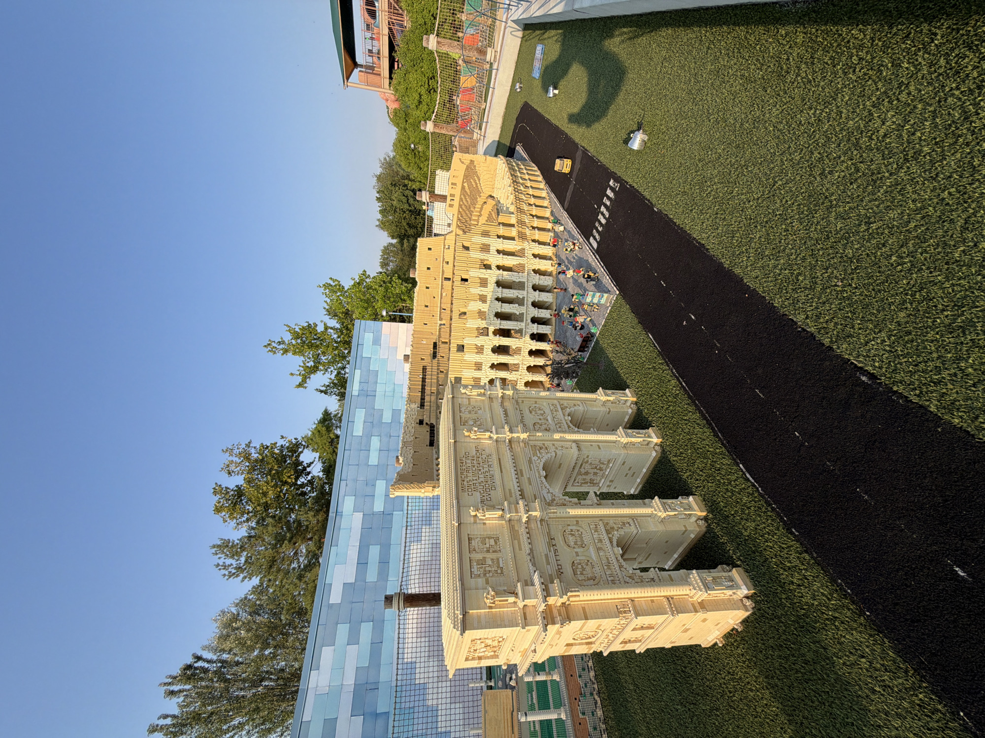 This image depicts a meticulously crafted Lego replica of the Roman Colosseum and Arch of Constantine, situated within the city of Castelnuovo del Garda, Italy. The scene is set against a clear blue sky, with a modern blue tiled wall and lush green artificial turf covering the ground. The Lego structures are detailed, showcasing the iconic architecture of both ancient Roman landmarks, complete with miniature figures populating the area around the Colosseum. A black Lego road with white crossing lines leads past the structures, and a miniature yellow vehicle is visible on the road. The overall environment suggests an outdoor attraction, possibly a theme park or a public display, designed to replicate historical Roman architecture using Lego bricks.