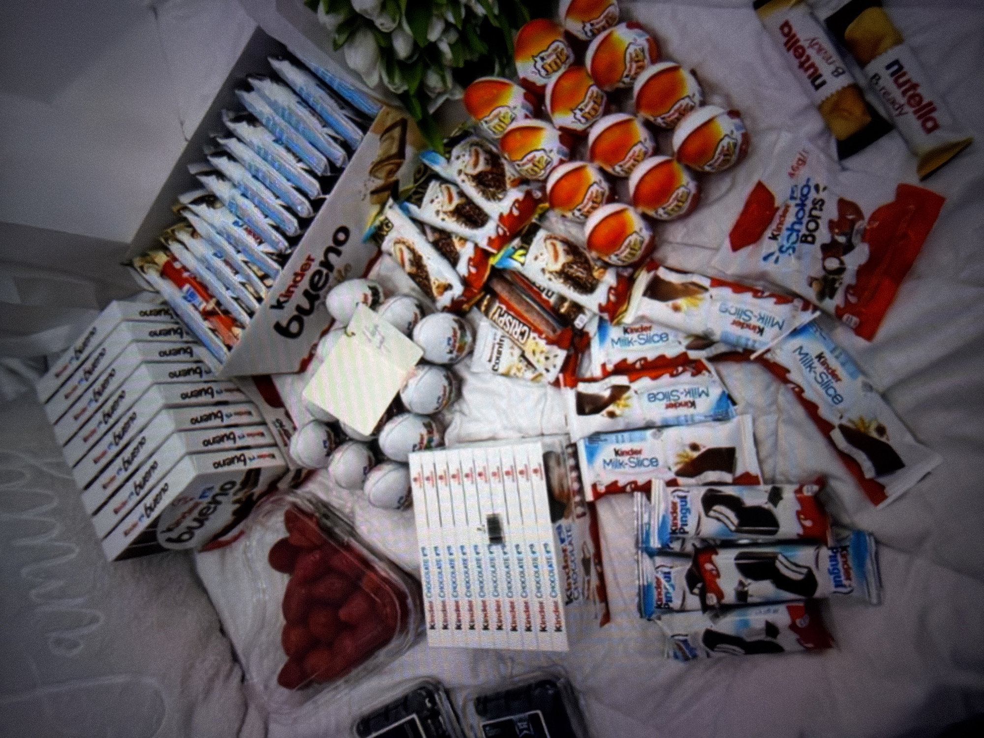 This is a high-angle, slightly out-of-focus photograph of a collection of Kinder brand chocolates and other sweets, as well as a container of raspberries, arranged on a white, textured surface. The arrangement appears to be on a bed, suggested by the soft, rumpled fabric and the presence of what looks like a throw pillow with the word "family" written on it in cursive.
Dominating the image are several boxes and packages of Kinder Bueno, Kinder Chocolate, Kinder Surprise eggs, Kinder Pingui, and Kinder Schoko-Bons. The Kinder Bueno boxes are stacked and also spread out, with one large box open, revealing multiple individually wrapped bars. There are also loose Kinder Surprise eggs, distinguished by their orange and white packaging. A small clear plastic container filled with red raspberries is visible in the upper left quadrant. In the lower right corner, two Nutella & Go snack packs are visible. A small bouquet of white tulips is also present.
The overall impression is a gift or a collection of treats. There are no people or explicit actions depicted. The lighting is soft, creating a slightly hazy effect. The location is identified as Beșalma, Moldova.