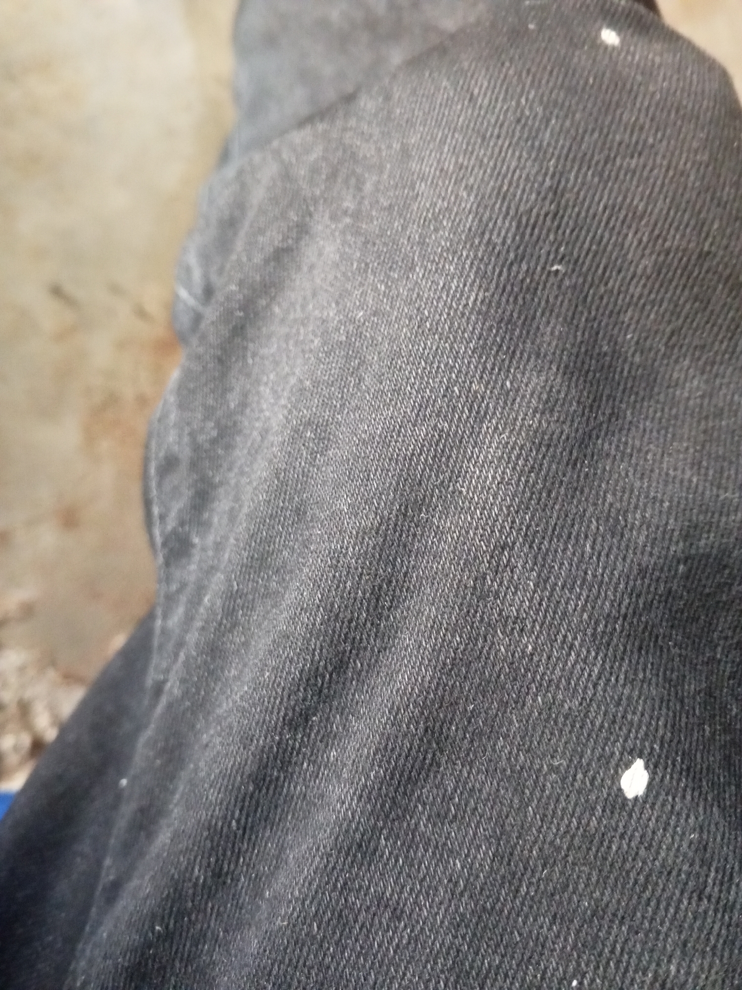 The image shows a close-up view of dark denim fabric. The fabric has a textured weave with diagonal lines. There are a few small white specks on the fabric, possibly paint or dust. The background is blurred and appears to be a neutral, earthy tone. The context of Nyabe, Nigeria, is not visually represented in the image.