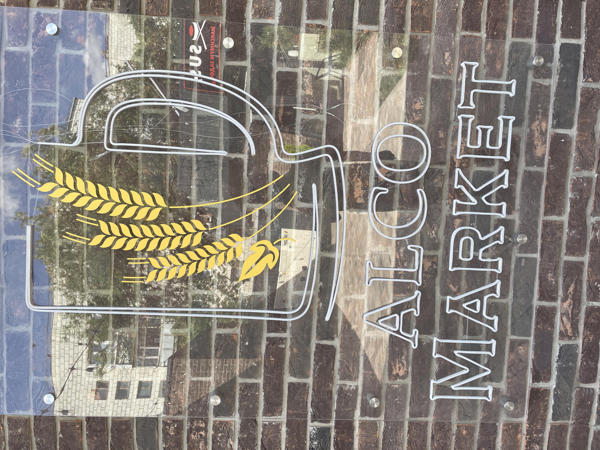 A clear glass sign is mounted on a brick wall. The sign displays a logo of wheat stalks within a beer mug outline, and below that, the words "ALCO MARKET" in white neon. The beer mug outline is made of white neon tubing. The wheat stalks are yellow. The bricks are dark brown and red. The background on the sign reflects a building and trees. There are four metal standoffs holding the sign to the wall.