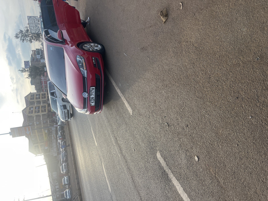 This image depicts an outdoor street scene, likely in Gitothua ward, Kenya, with a strong focus on a red Volkswagen Golf car. The car is prominently parked on an asphalt surface, angled towards the right of the frame. Its front license plate, "KDN 797W," is clearly visible, along with the Volkswagen emblem. One of the car's doors is open, and a person's legs, clad in light trousers and dark shoes, are partially visible near the opening, suggesting someone is either entering or exiting the vehicle.
In the background and extending along the left side of the frame, several other cars and a white van are parked in a line. Beyond these vehicles, multi-story buildings with varied facades line what appears to be a street, contributing to an urban or semi-urban setting. The sky is bright on the left, indicating either early morning or late afternoon, with the sun creating a strong glare and suggesting clear weather. The ground is paved with asphalt, featuring white painted lines that denote parking spots or road boundaries. Scattered small pieces of debris, including a dried leaf, are visible on the asphalt. The photograph is taken with a tilted horizon.