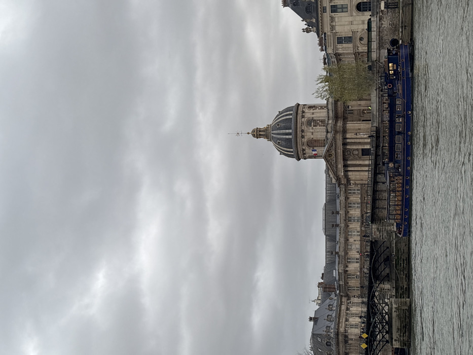 The image presents a daytime view of the Seine River in Paris, France, facing the north bank under an overcast sky. The central feature is the Institut de France, identifiable by its prominent grey dome topped with a spired weathervane and a small French flag. The building displays classical architecture with stone facades, columns, and arched windows, flanked by other historic Parisian structures featuring grey slate roofs. To the left of the Institut, a section of the Pont des Arts, a pedestrian bridge with dark metal arches and stone piers, extends across the river; several human figures are indistinctly visible walking on its surface. A long, dark blue passenger boat, equipped with numerous windows, is positioned in the foreground on the river, either moored or slowly navigating parallel to the bank. The sky is uniformly covered with thick, grey clouds, and the river water exhibits a dark, rippled surface. Sparse, light green foliage is visible along the river's edge and between buildings.