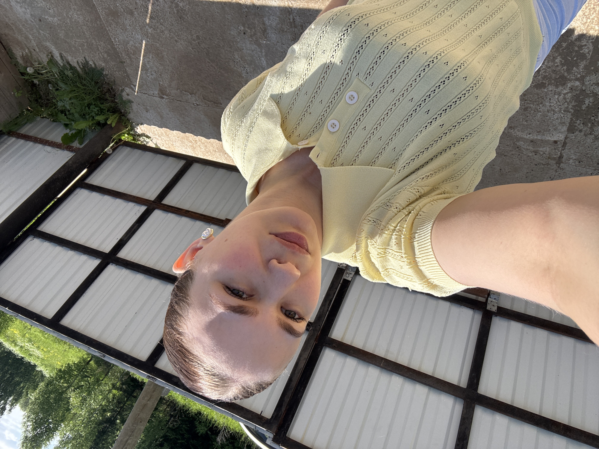 A young woman is depicted in a close-up, high-angle selfie taken outdoors in Nizhnyaya Gar', Russia. She has light skin, dark eyebrows, and her dark hair is slicked back. She looks directly at the camera with a neutral expression. She wears small, ornate earrings, possibly white pearl or gemstone with a gold-tone outline, in both earlobes. Her attire consists of a light yellow, short-sleeved collared polo shirt featuring an openwork knit pattern with vertical striped details and small eyelets. The shirt has a three-button placket with white buttons. A portion of light-colored lower garments, likely white or light blue trousers, is visible at the bottom left. The background features a white corrugated metal roof structure supported by dark brown or black beams, which appears to be a shed or an outbuilding. Behind this structure, green trees and foliage are visible under a bright blue sky with sparse clouds, indicative of a sunny day. The ground in the foreground and beneath the structure is concrete with visible cracks and patches of weeds, including dandelions, growing at the base of the structure. Direct sunlight casts shadows across the scene.