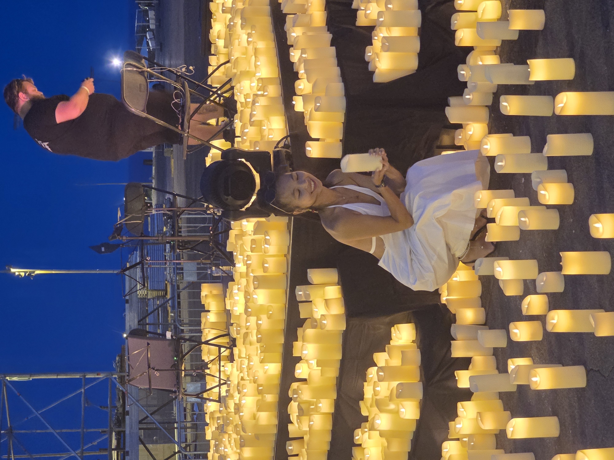 A large outdoor field is illuminated by numerous lit candles arranged in rows and on raised platforms, creating a warm, glowing ambiance against a dark blue night sky. In the foreground, a young woman wearing a white dress is kneeling and smiling, holding a single candle in her hands. She is surrounded by a dense field of lit candles, some of which are placed on dark fabric-covered risers. In the mid-ground, a man with a beard, wearing a black t-shirt and dark pants, stands on a folding chair. He appears to be interacting with something in his hands, possibly preparing or adjusting candles. Behind him, scaffolding and stage equipment are visible, suggesting a performance or event setup. The location is identified as the Parc del Fòrum in Sant Adrià de Besòs, Spain.