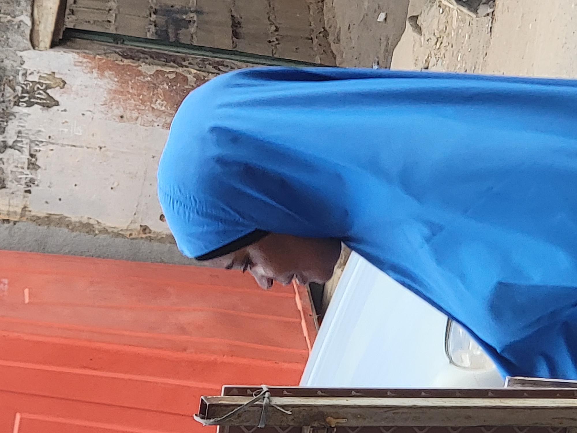 A person wearing a blue covering is seen from the side, partially obscured by the material. Below the person is a white surface, and below that is an orange surface with horizontal grooves. A metal frame with wire ties is at the bottom. The background includes a wall with visible wear and tear. The image context is Maiburiji, Nigeria.