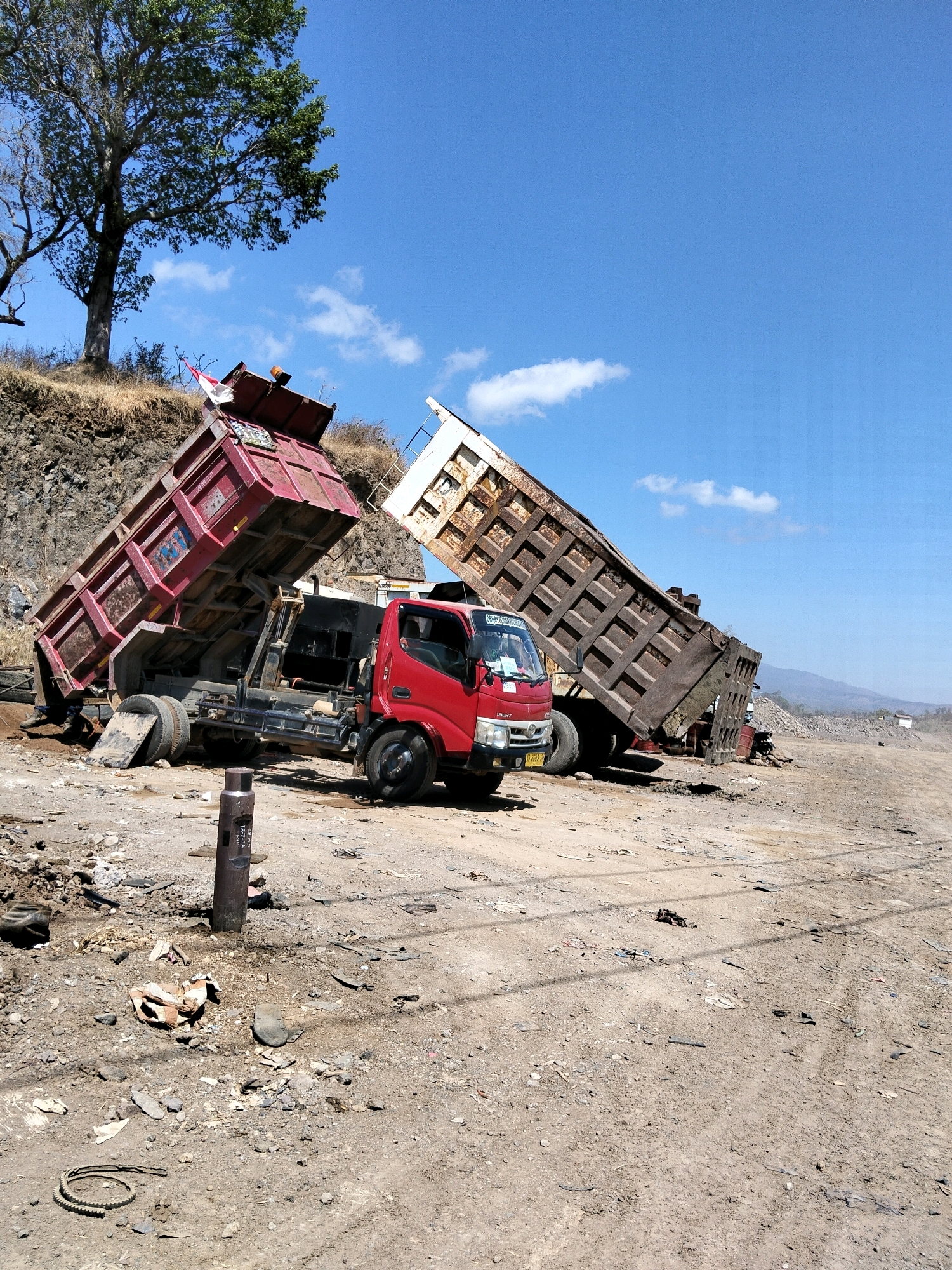 In Bujel, Indonesia, a red dump truck is positioned on a dirt lot with its cargo bed raised. Another dump truck, older and rusted, has its bed also raised and appears to be partially tipped over. The surrounding environment consists of dry earth, scattered debris, and a rocky embankment with sparse vegetation and a large tree. The sky is clear and blue with a few scattered clouds. The scene appears to be an outdoor industrial or construction area.