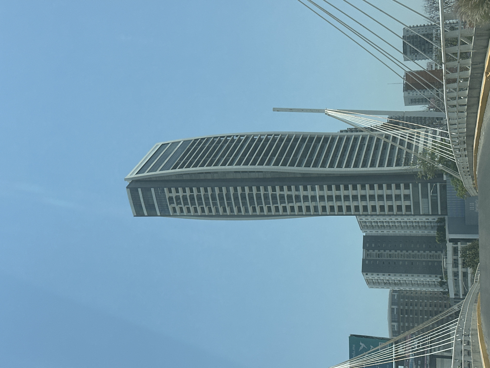 This image depicts a modern urban landscape in San Bernardino Tlaxcalancingo, Mexico, under a clear, bright blue sky during the daytime. The central subject is a tall, striking high-rise building with a distinctive, gently curving facade. The upper section of this building features numerous vertical slats or window frames, while the lower part displays a grid pattern of rectangular windows.
To the right of the main building, a section of a cable-stayed bridge or overpass is prominently visible, characterized by multiple white cables extending from a central support (partially out of frame) to the bridge deck. The roadway of this bridge appears to be asphalt. In the background, several other multi-story buildings, varying in height and design, contribute to the city skyline. One building in the distance has a reddish-orange top, possibly indicating ongoing construction or a unique architectural feature.
The scene appears static, with no people or explicit activities visible. The photograph is likely taken from a vehicle or vantage point on the bridge, offering a view of the modern architecture against the expansive blue sky. A green billboard in the bottom left corner, partially obscured, shows legible text "edu.mx". The overall impression is one of a developed, contemporary urban environment.