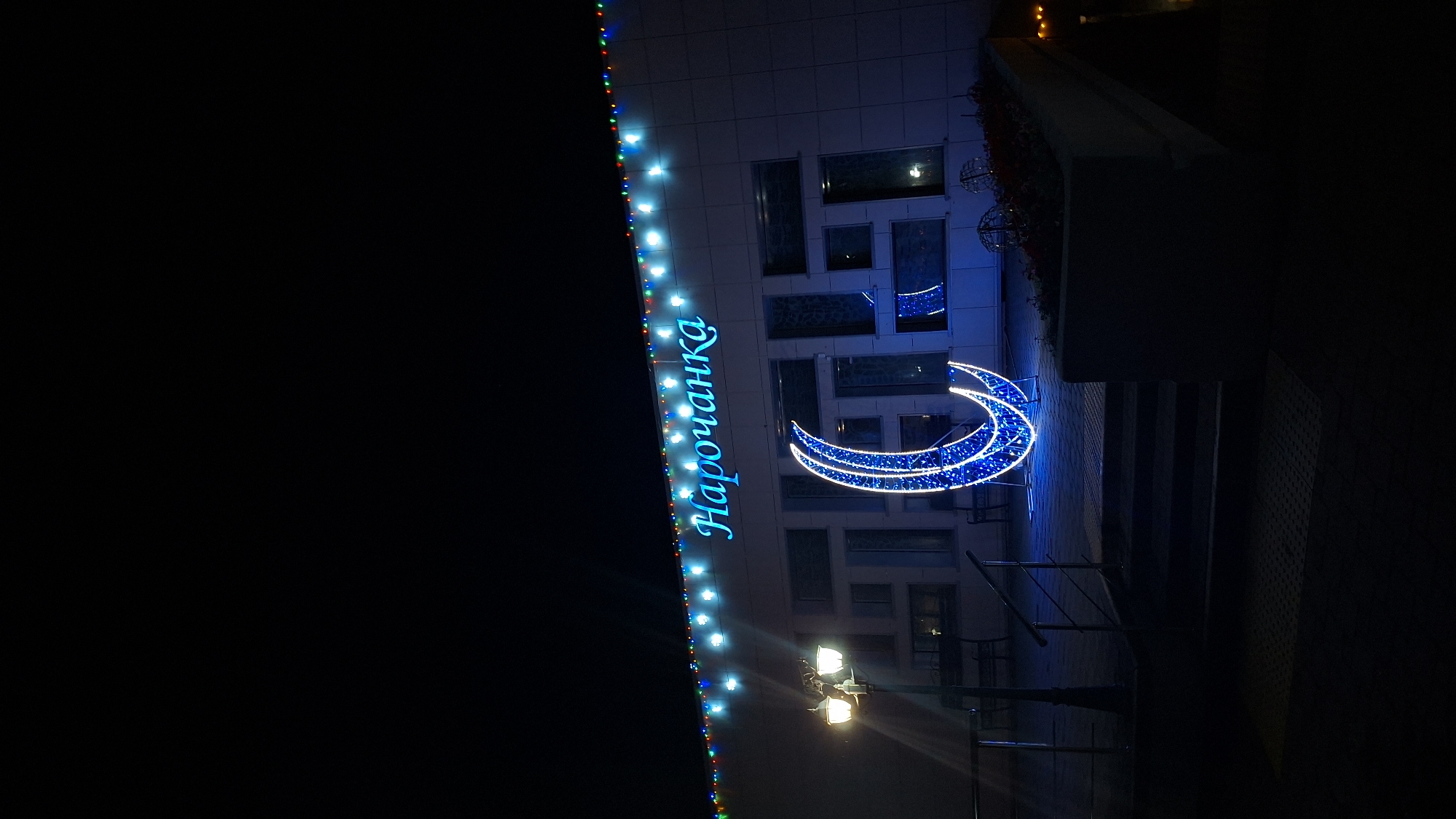 At night in Нарацкі сельскі Савет, Belarus, a building is decorated with blue lights outlining the word "Нарочанка" and a crescent moon. The building has a modern facade with rectangular windows. String lights run along the building's edge. A streetlight illuminates the area. Stairs are visible to the right of the illuminated crescent. The background is dark.