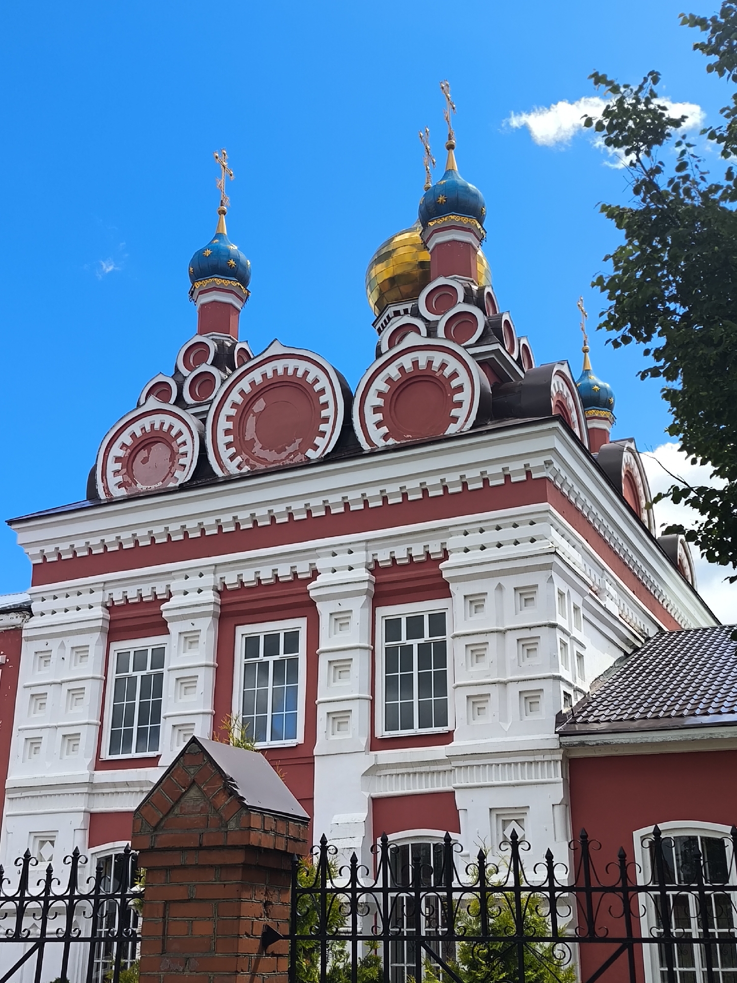 The image shows a side view of a red and white Orthodox church with blue and gold domes against a bright blue sky with some clouds. The church has ornate architectural details, including decorative white trim and arched windows. In the foreground, there is a black wrought-iron fence in front of a brick pillar, and some greenery. The overall scene suggests a sunny day in a town or city in Russia, likely Taldom, given the architectural style. There are no people visible in the image.