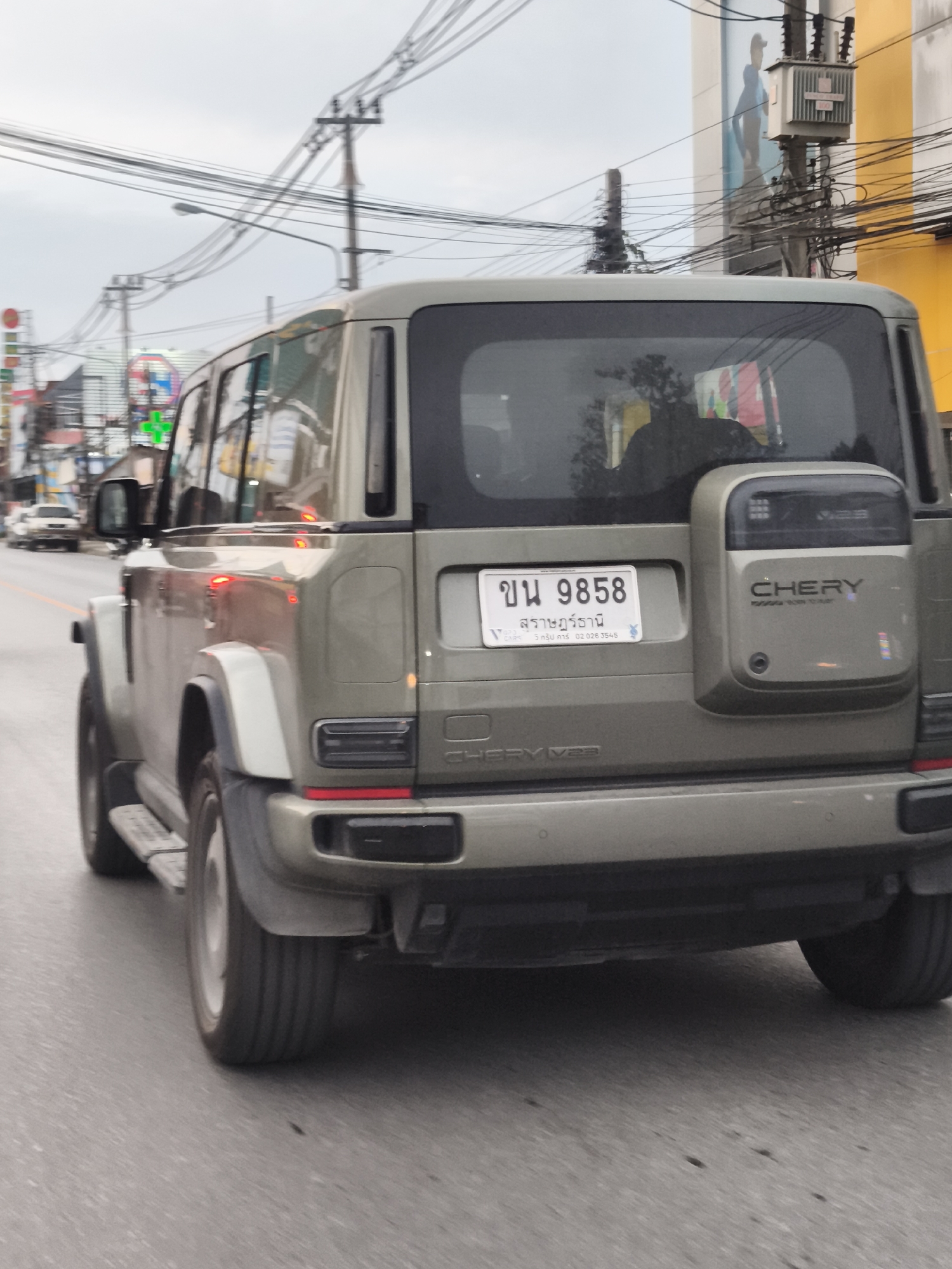 The image shows the rear of a grey Chery vehicle driving on a street in the Ko Samui District of Thailand. The vehicle has a license plate that reads "ข น 9858" with Thai text below it. A spare tire is mounted on the rear of the vehicle. The background shows a street scene with power lines, traffic lights, and buildings. The sky is overcast.